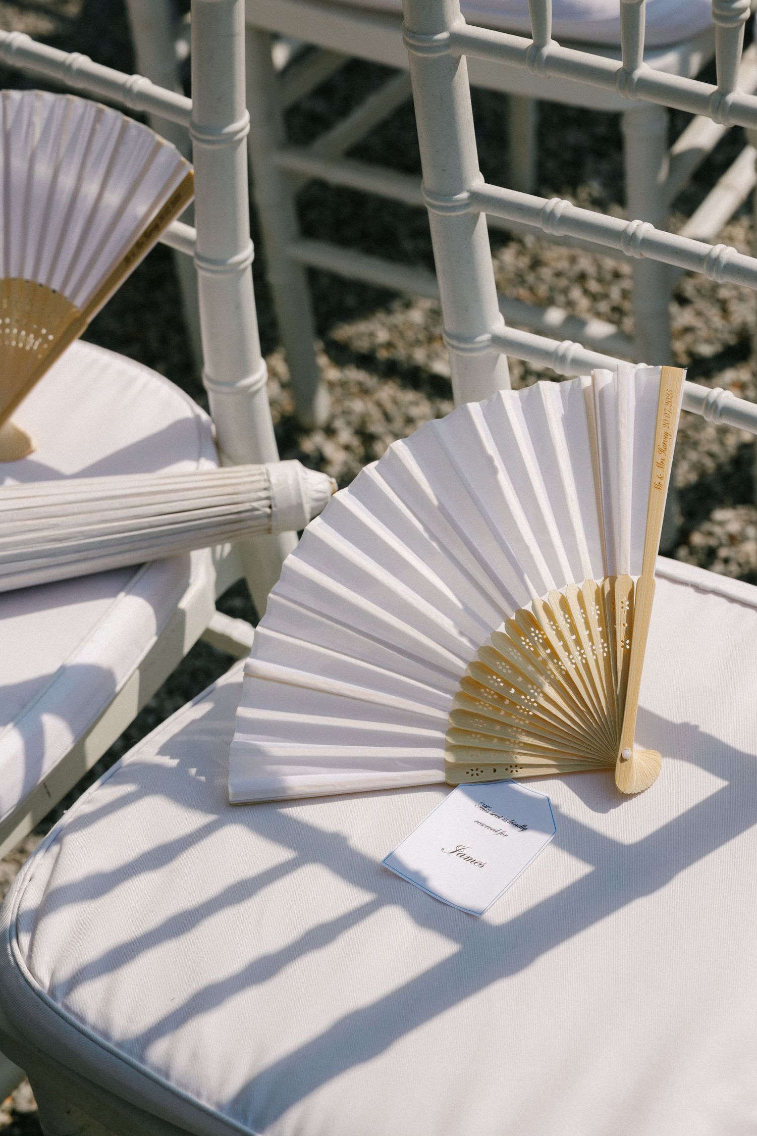White and gold folding hand fans placed on white cushioned chairs at an outdoor wedding ceremony.