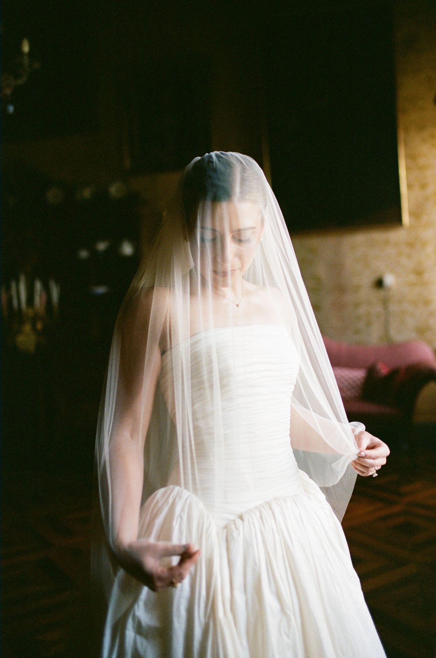 Bride in strapless white wedding gown and sheer veil looks down elegantly in a softly lit vintage room.