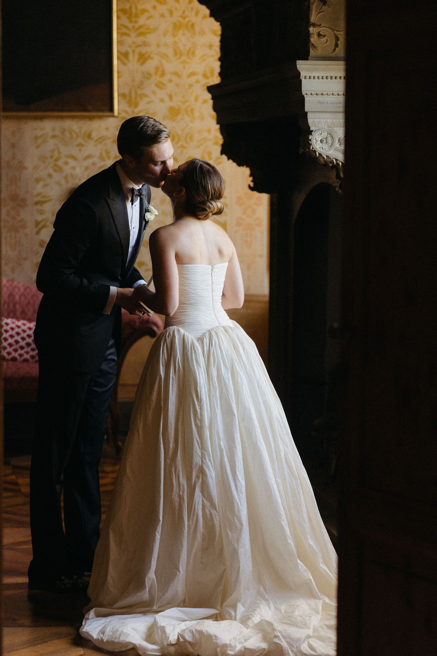 Groom in black tuxedo kissing bride in strapless ball gown in ornate wedding venue.