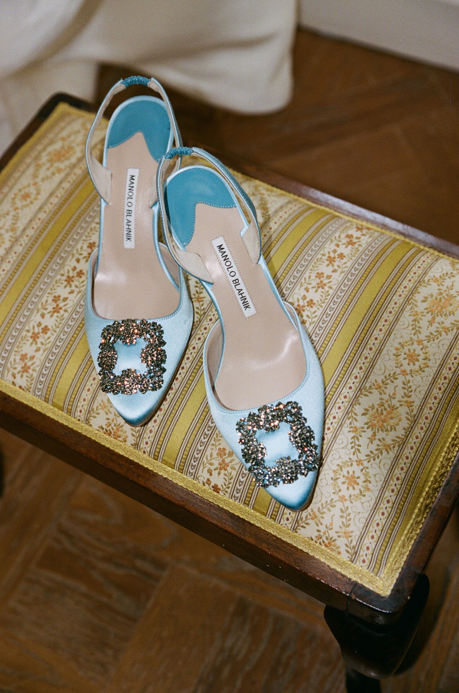 Light blue Manolo Blahnik slingback heels with jeweled buckles displayed on a vintage upholstered stool.