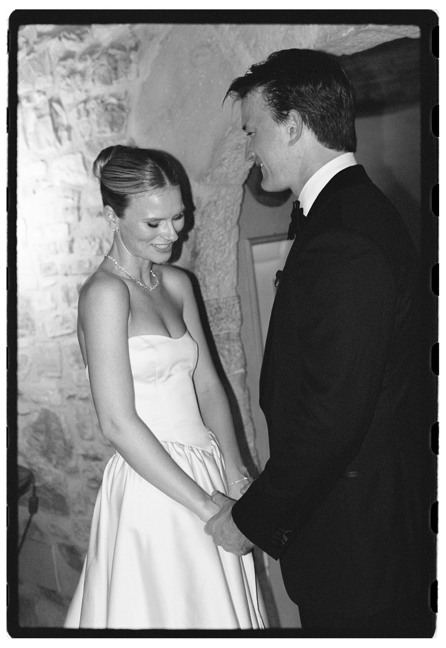 Black and white wedding photo of bride in strapless gown and groom in tuxedo holding hands and smiling.