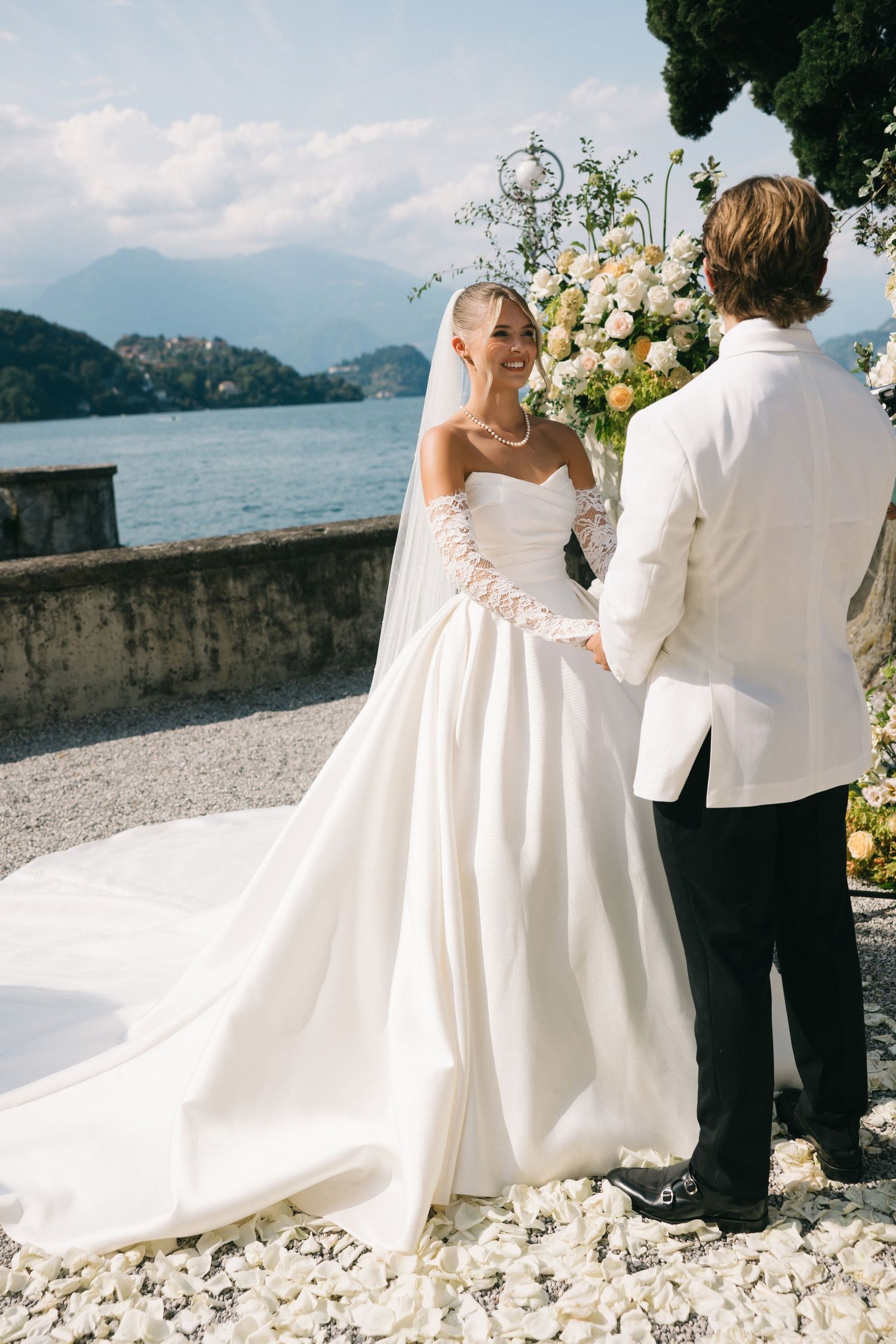 Bride in white strapless gown with veil holds hands with groom in white jacket at outdoor lakeside wedding ceremony.