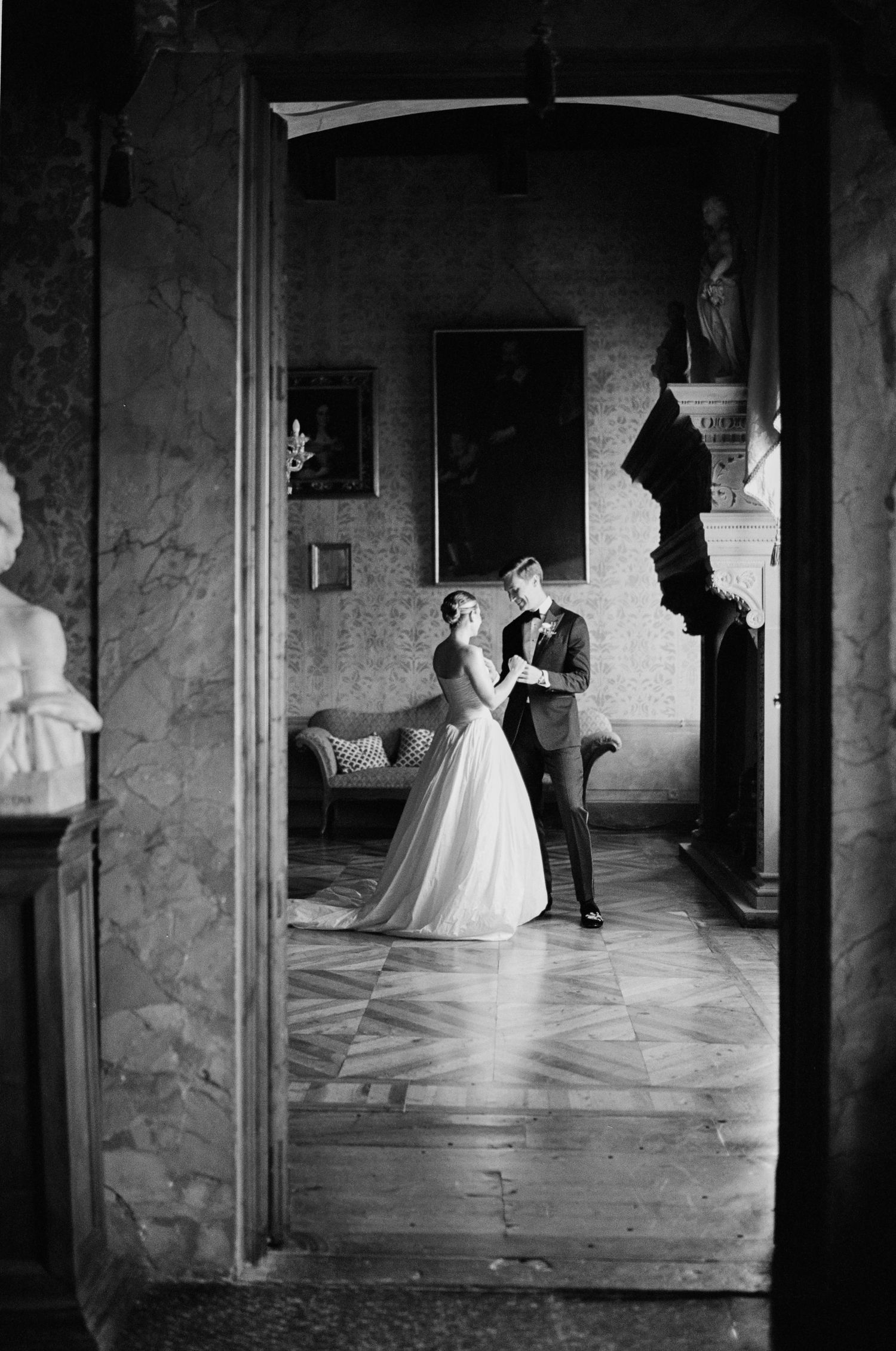 Black and white photo of bride and groom dancing in grand classical villa framed by marble doorway.