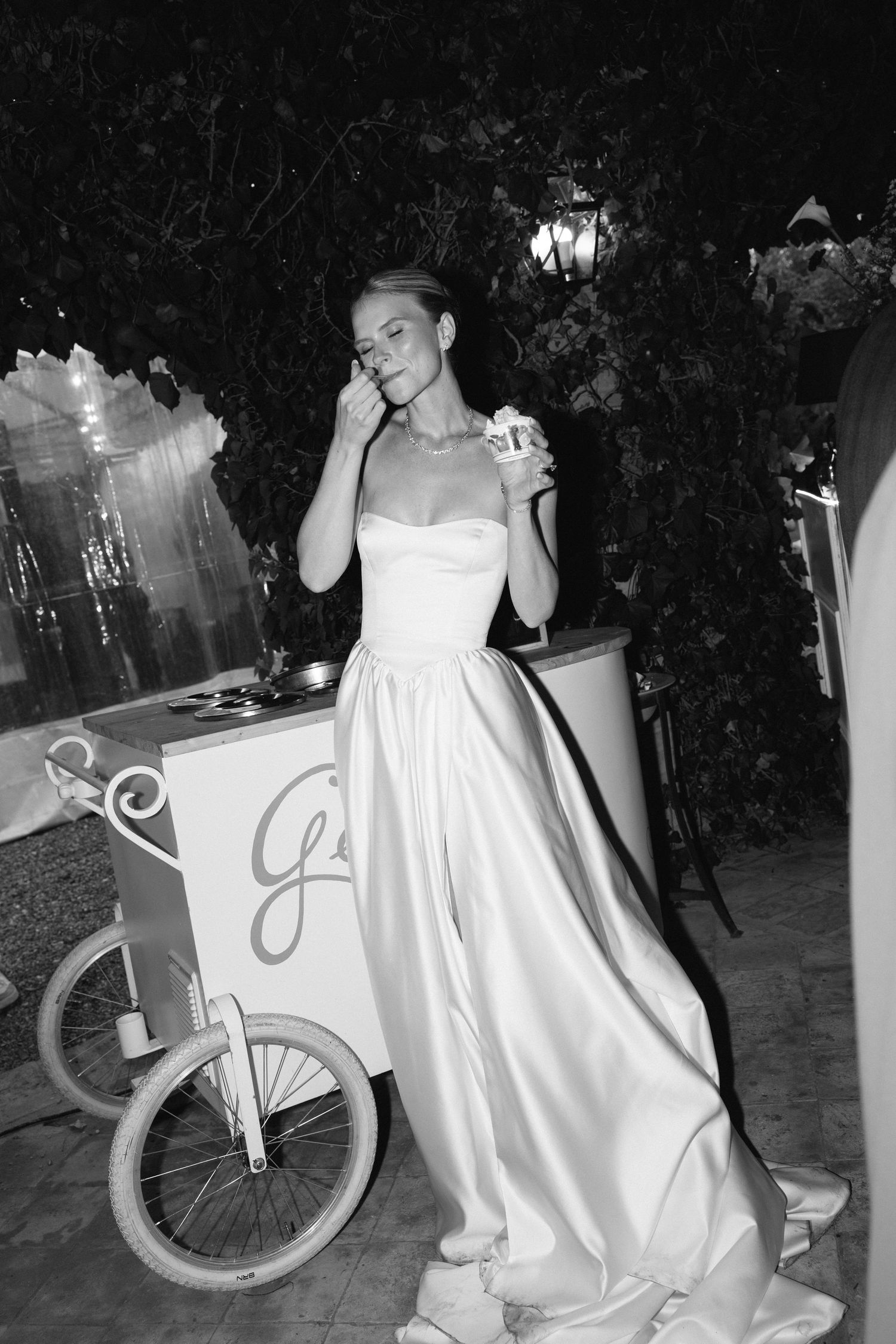 Black and white photo of a bride in a strapless satin gown holding a treat near a vintage dessert cart outdoors.