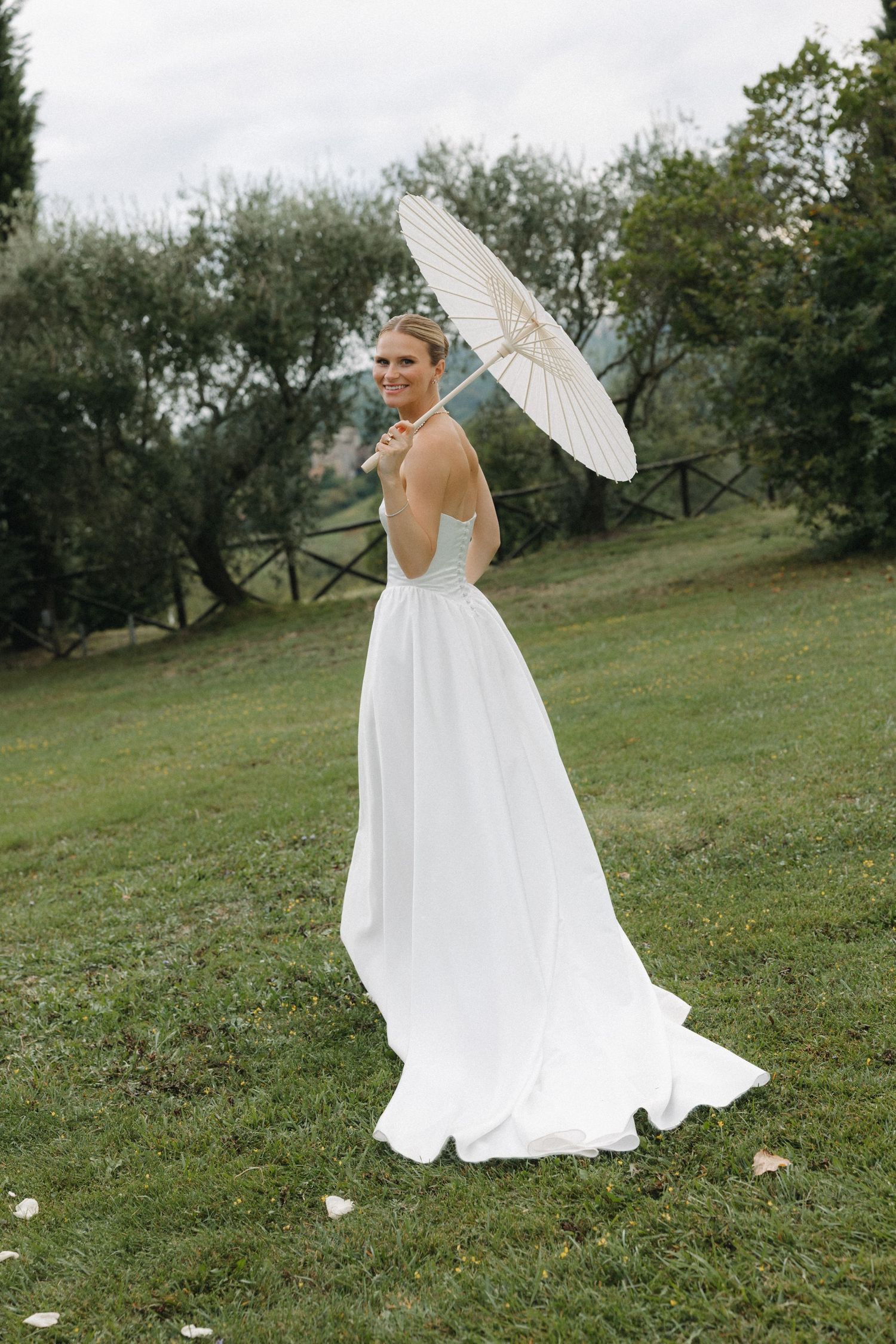 Bride in backless white gown holding parasol umbrella on green lawn with trees in background.