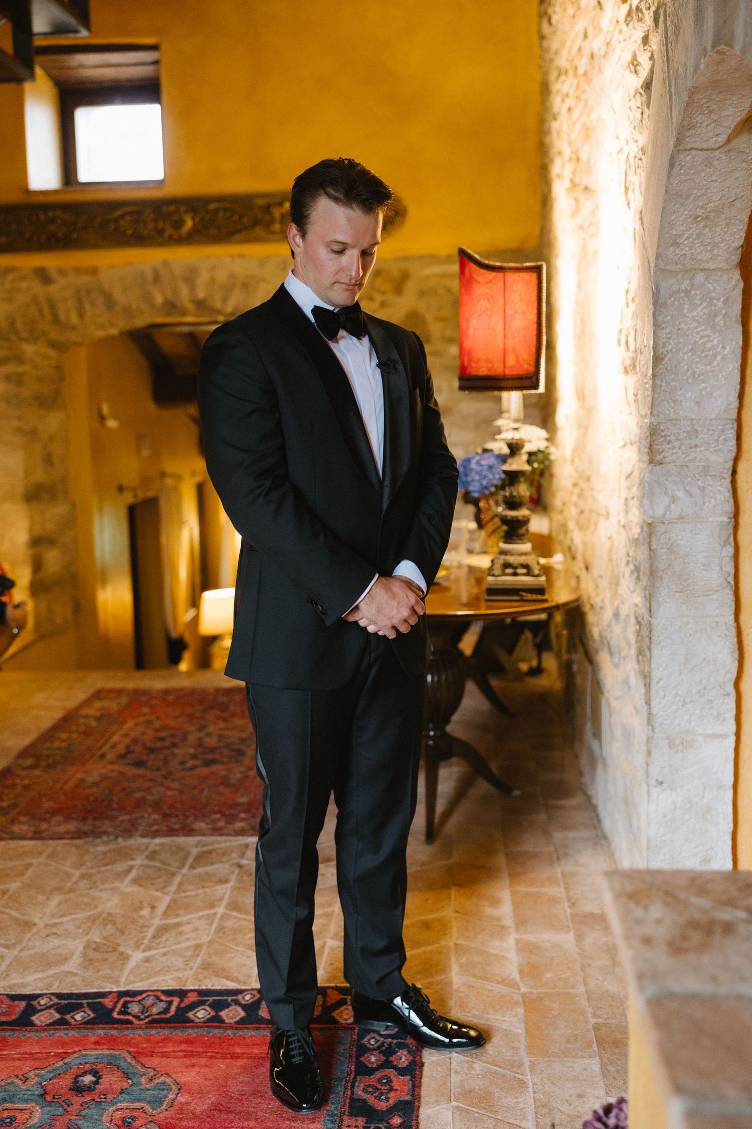 A groom in a black tuxedo with bow tie stands thoughtfully in a rustic stone-walled venue with warm lighting.