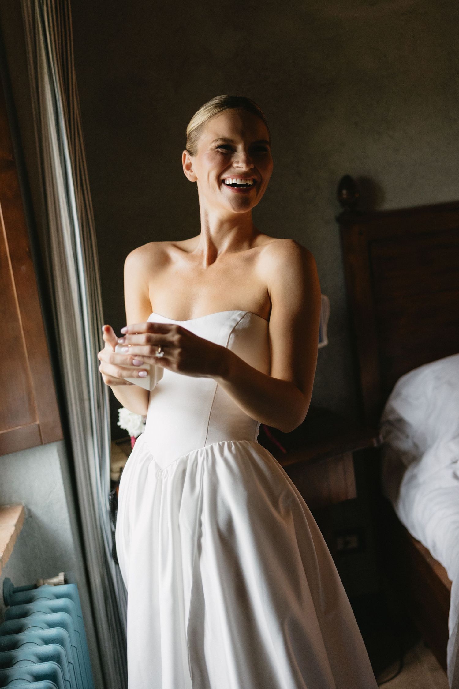 Smiling bride in strapless white wedding gown holding a glass, standing near a window in an elegant bedroom.