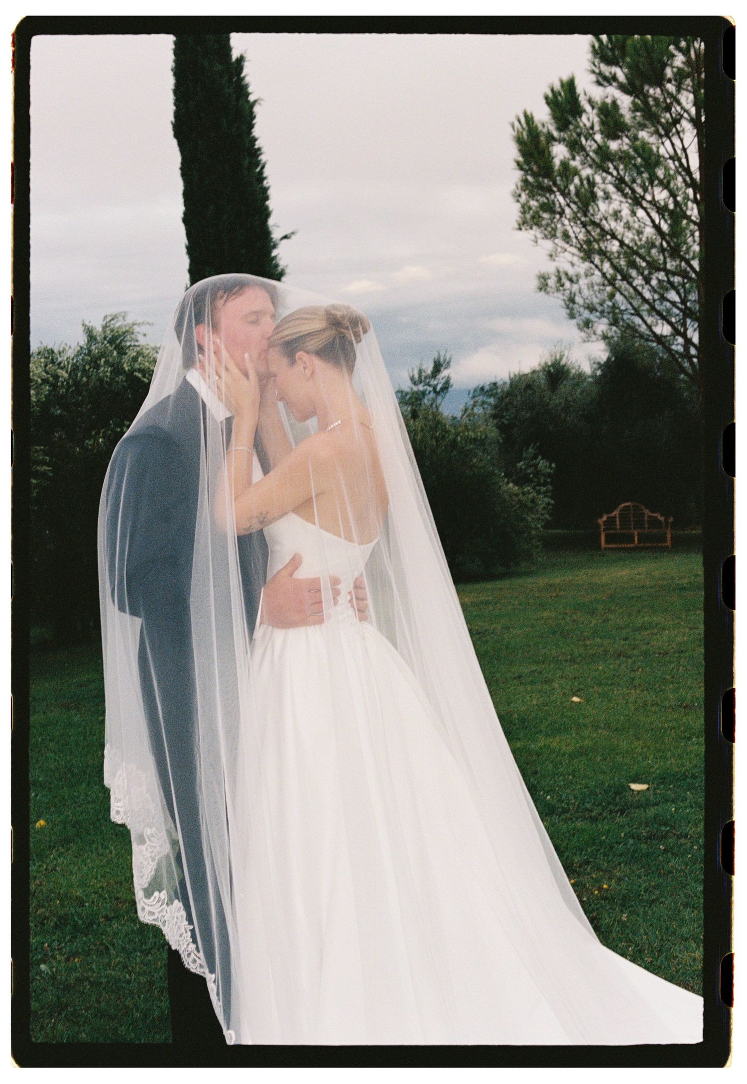 A bride and groom share a romantic kiss under a flowing white veil outdoors on lush green grounds.
