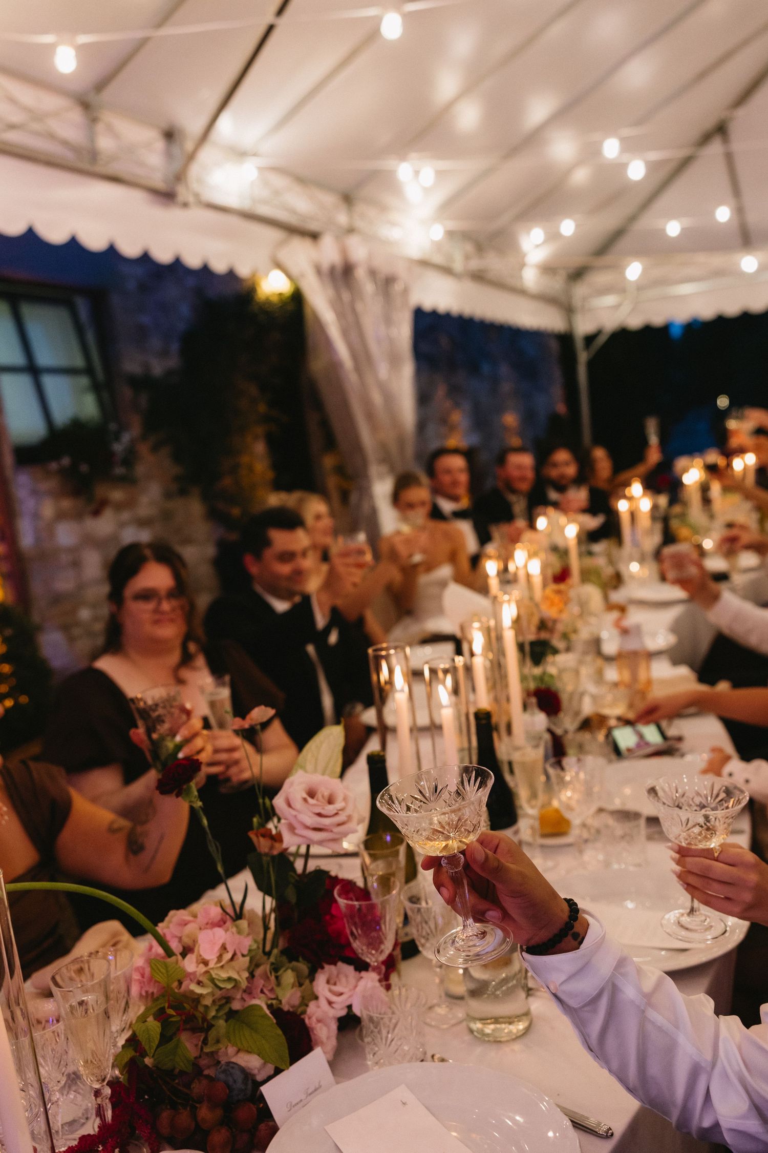 Elegant wedding reception under a tent with guests toasting, candles, floral centerpieces, and string lights at dusk.