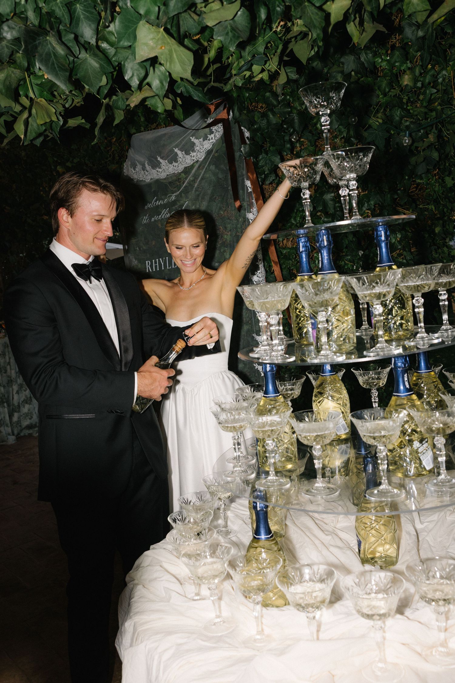Bride and groom pouring champagne tower at wedding reception with crystal glasses and lush greenery backdrop.