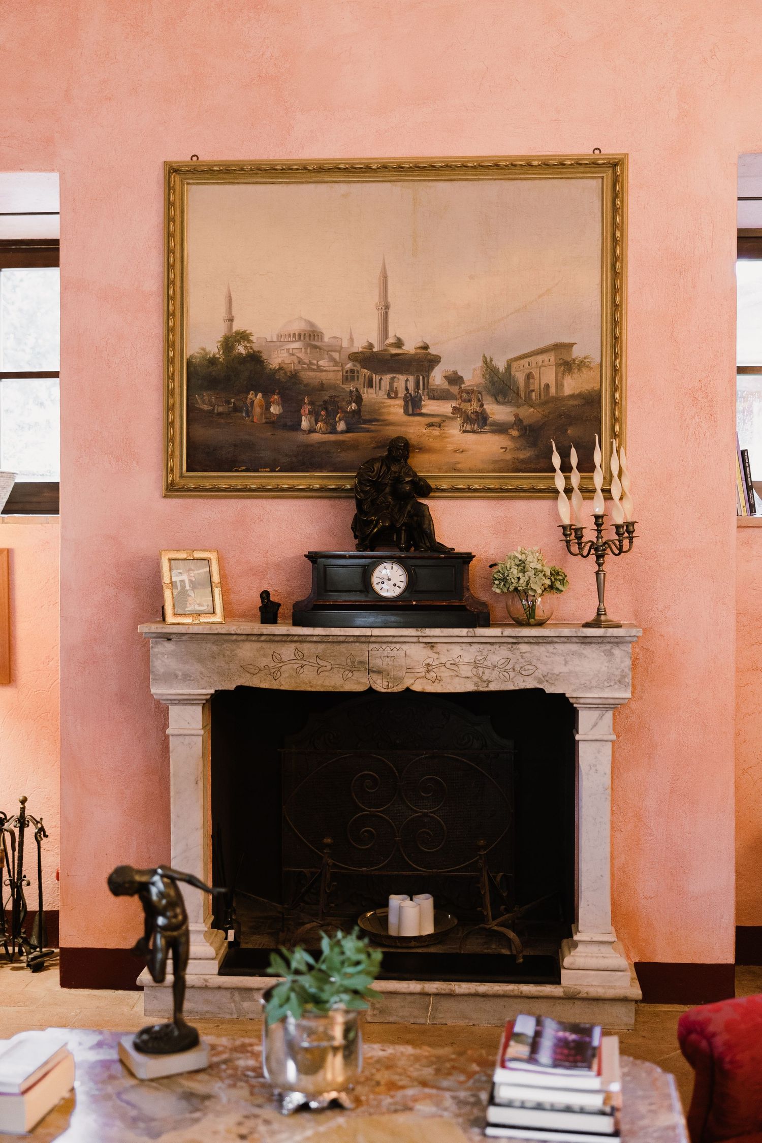 Elegant fireplace with marble surround, black clock, candelabra, and vintage landscape painting on pink wall.
