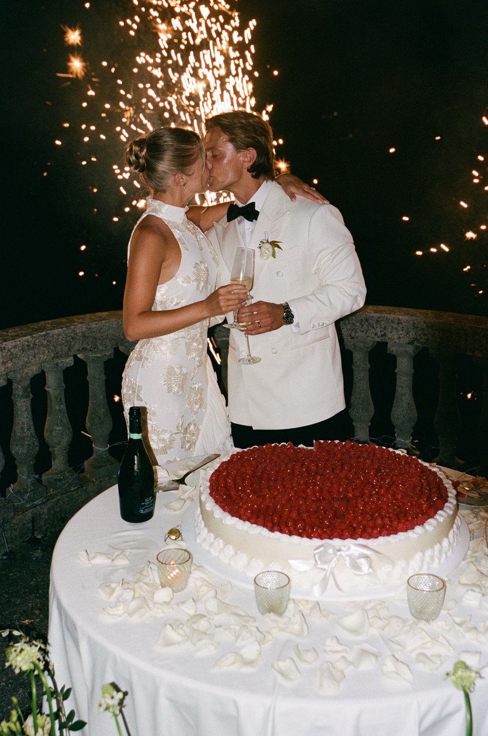 Newlyweds kissing by wedding cake with sparklers at night reception celebration.