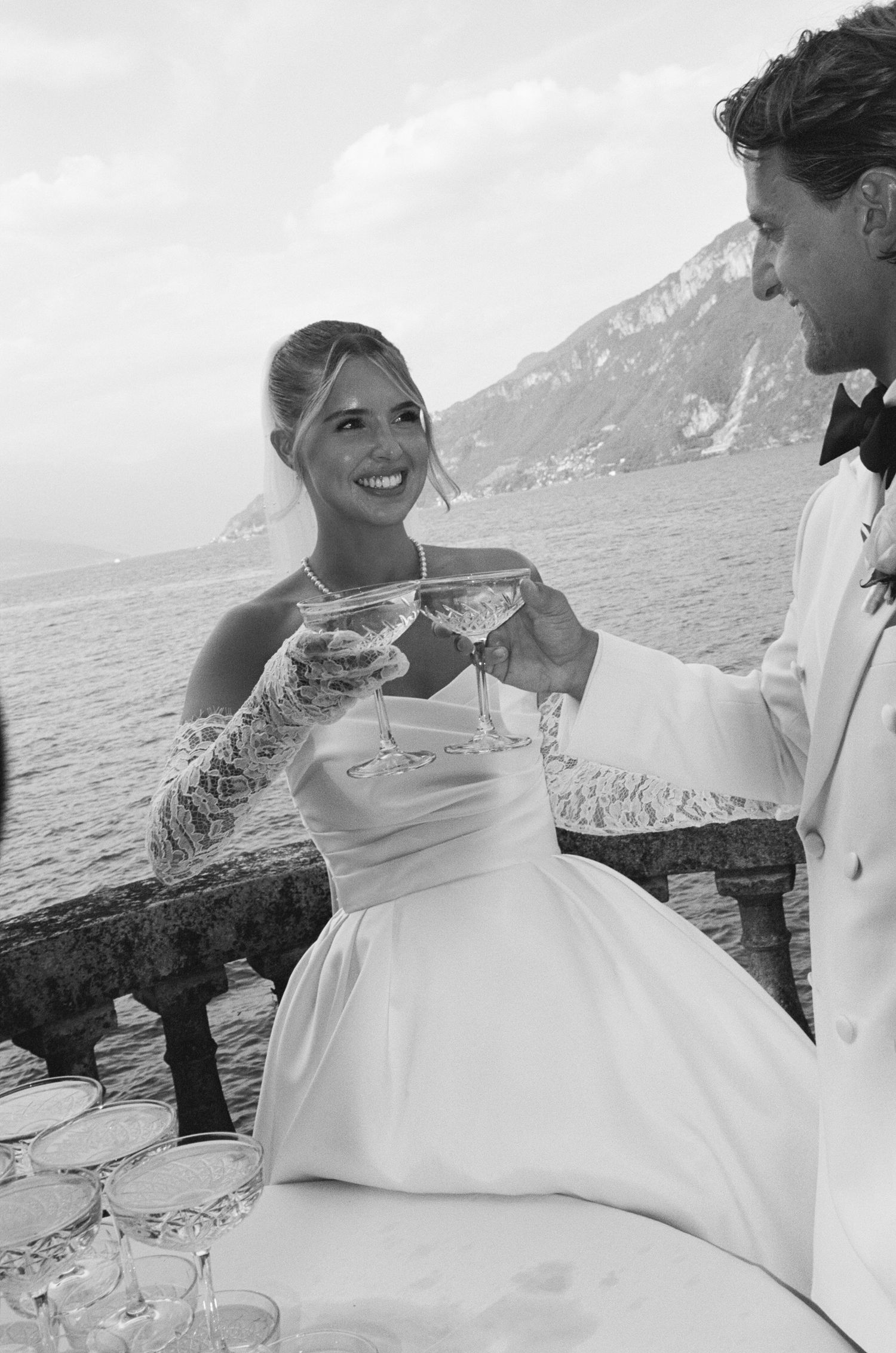 A smiling bride in a white gown toasts champagne with a groom in a white suit, with a scenic coastal backdrop.