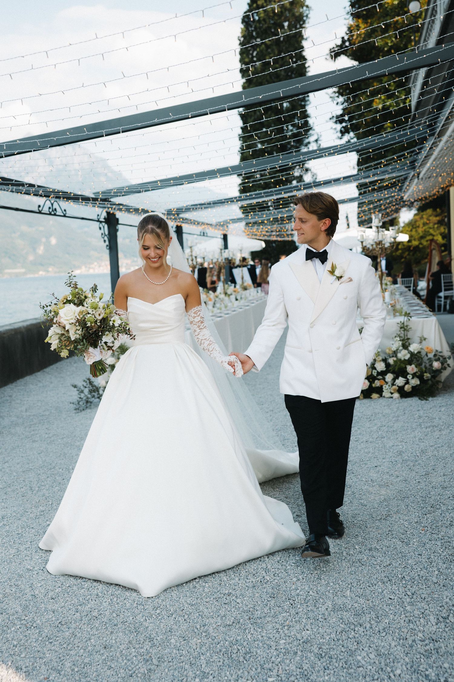 Bride in strapless white ball gown and groom in white tuxedo hold hands at outdoor lakeside wedding reception.