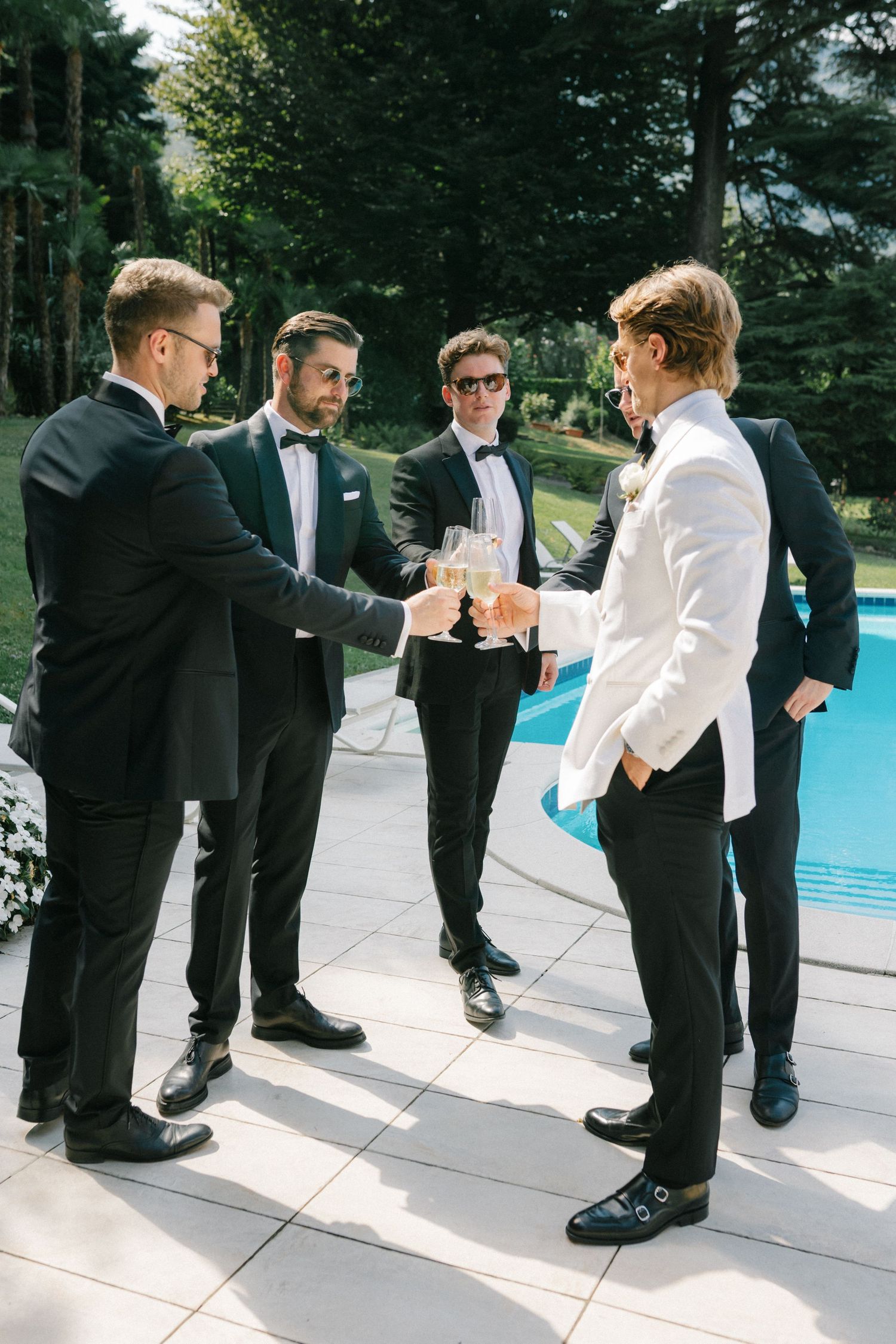 Groom and groomsmen in tuxedos toasting with champagne glasses by a pool at an outdoor wedding.