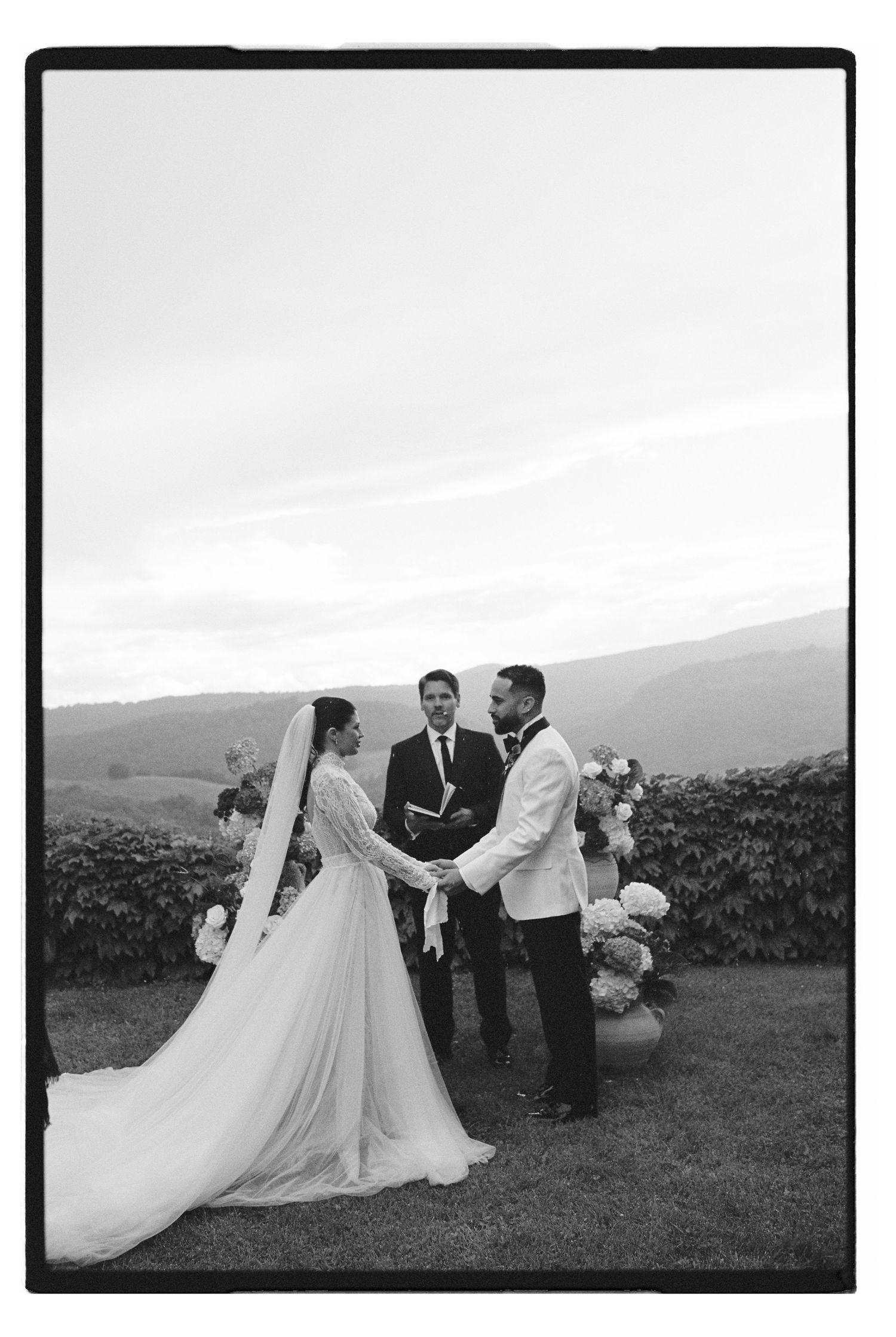 Black and white outdoor wedding ceremony with bride in flowing gown, groom in white jacket, and officiant against mountain backdrop.