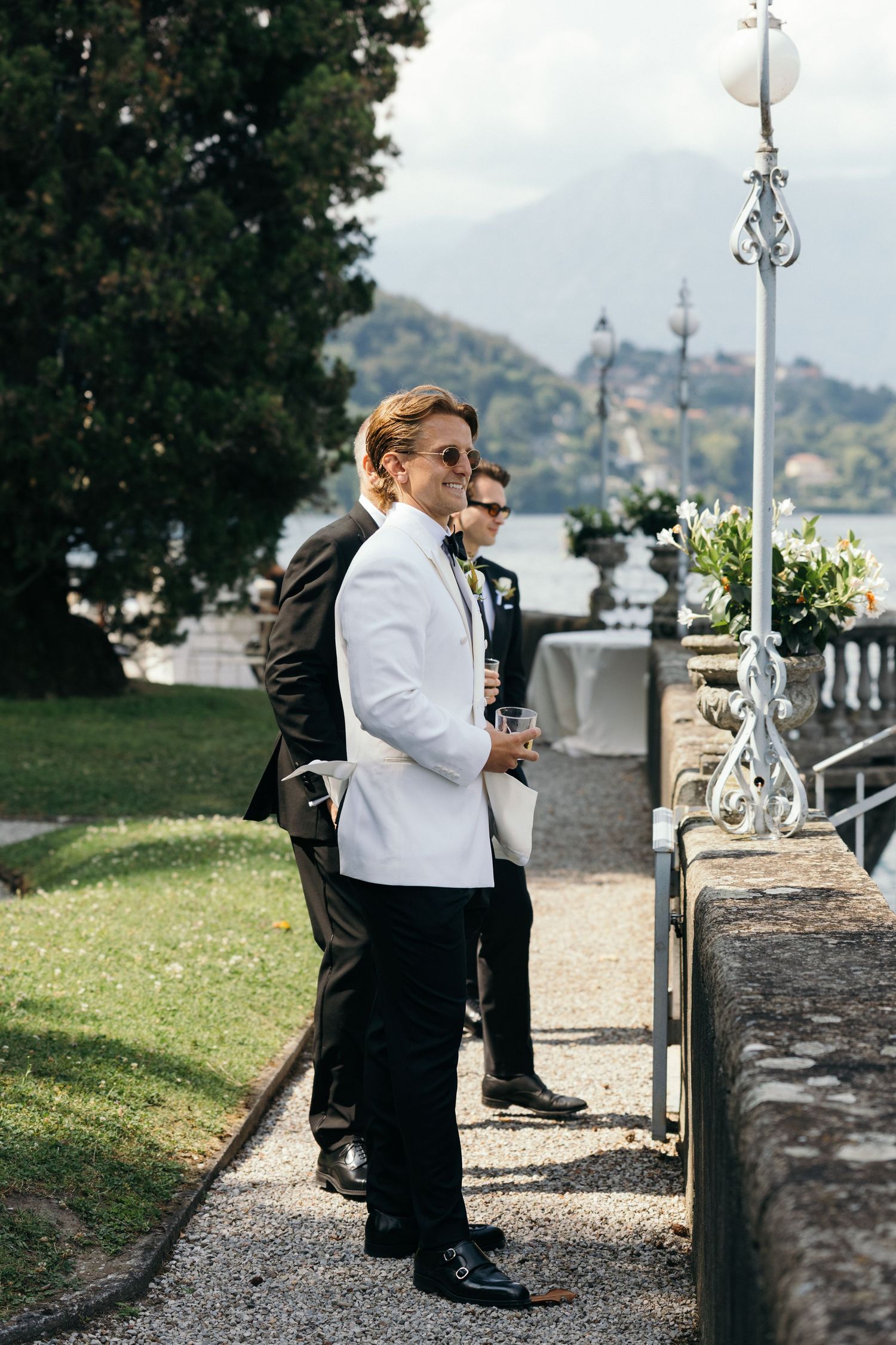 Groom in white tuxedo jacket holds drink outdoors at elegant lakeside wedding venue with mountains in background.