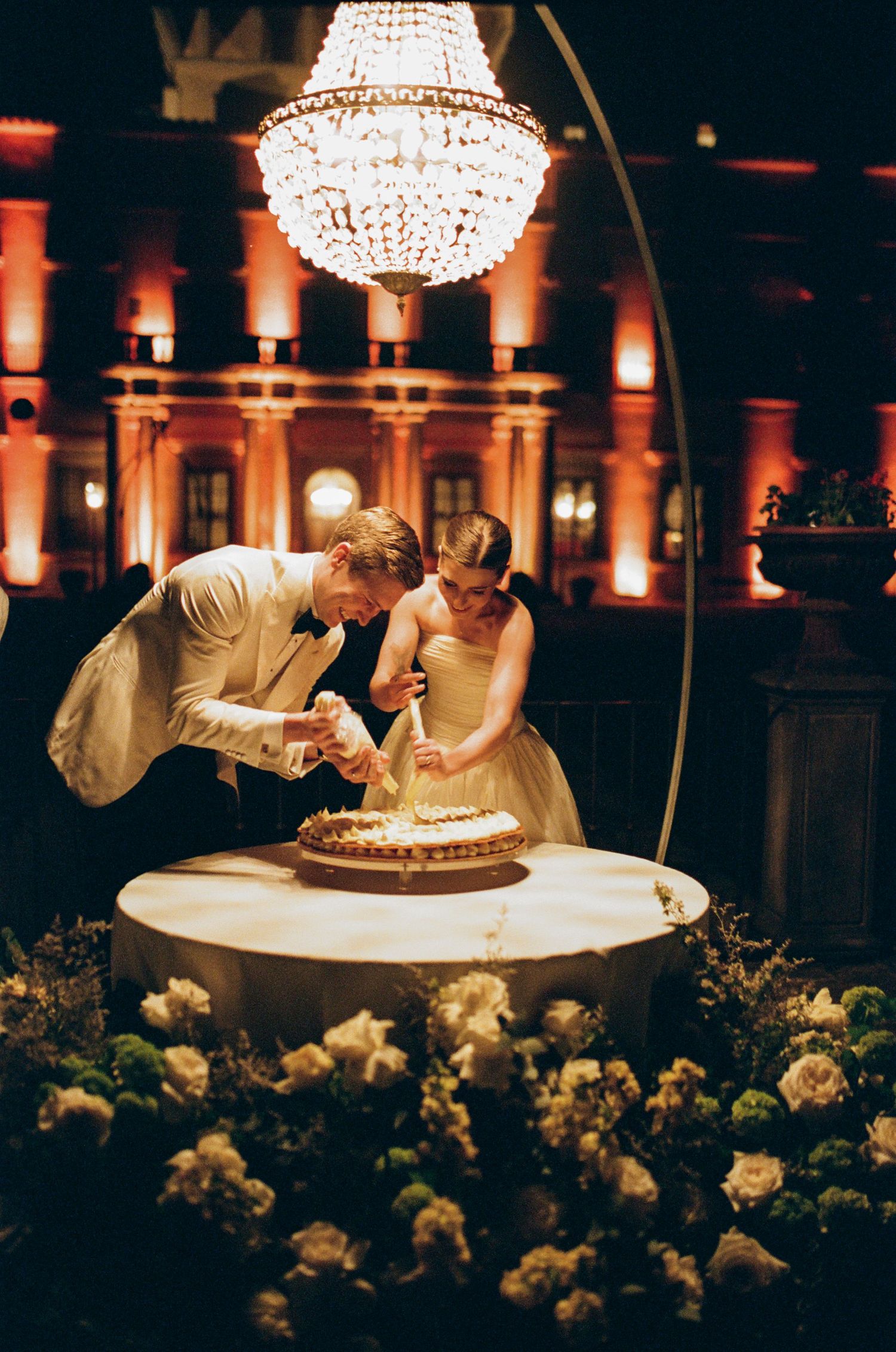 Bride and groom cutting wedding cake at elegant evening reception with chandelier and warm amber uplighting.