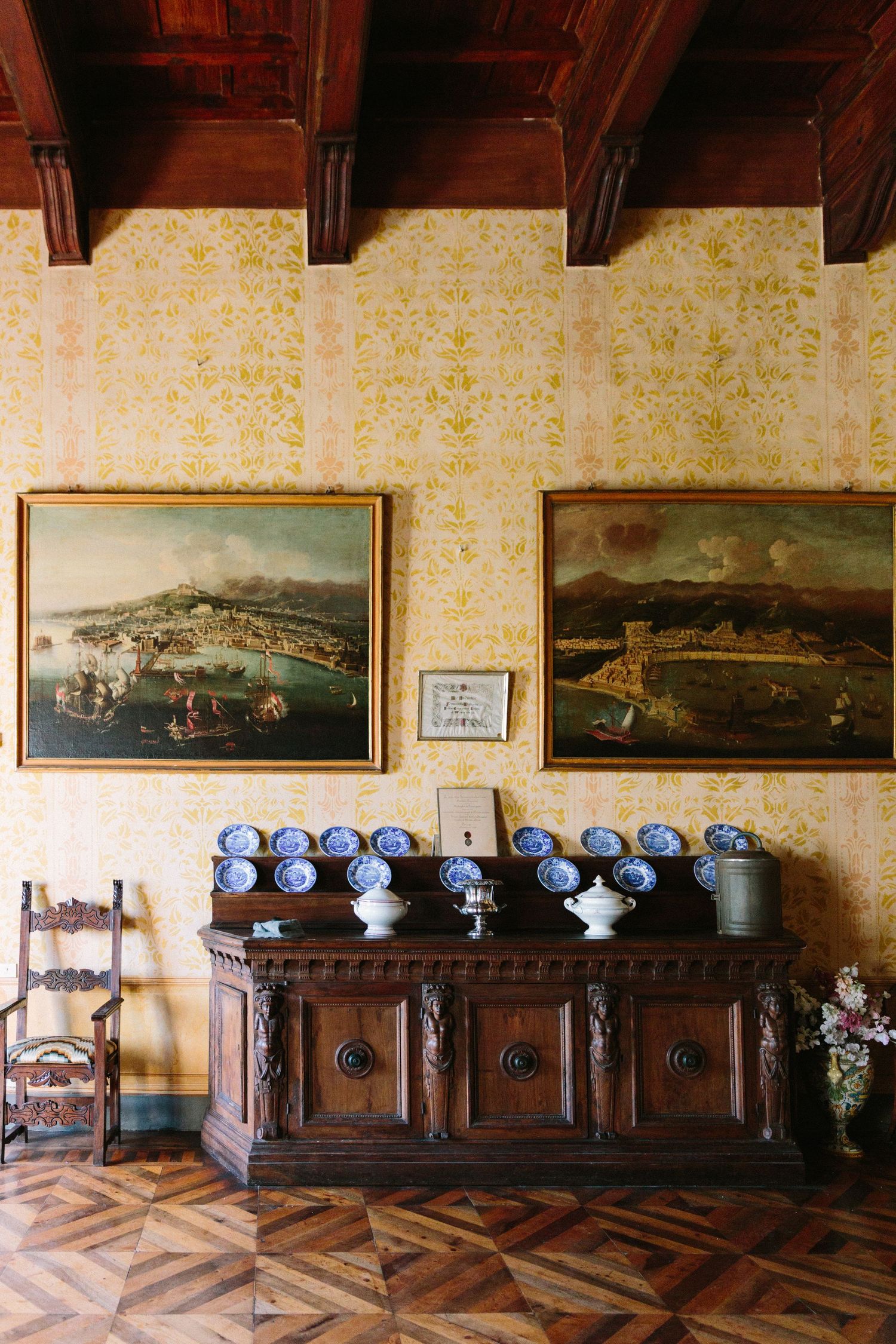 Ornate wooden sideboard displaying blue and white porcelain beneath two large landscape paintings on yellow damask wallpaper.