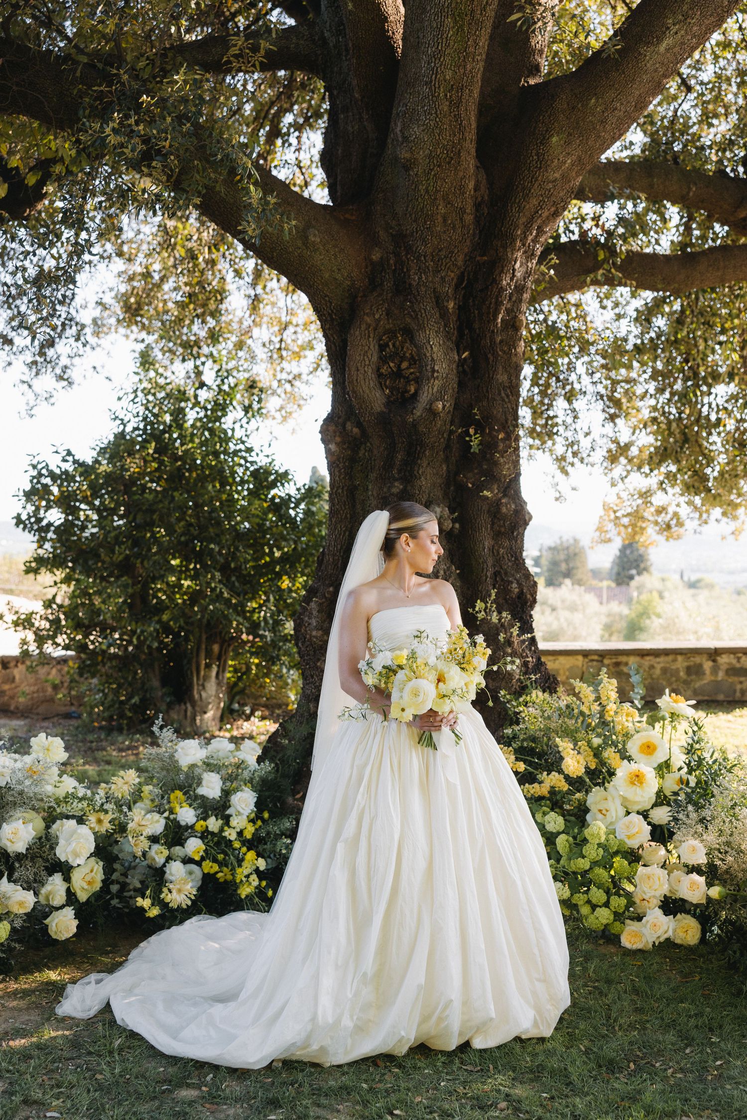 Bride in full white gown with veil holds yellow bouquet under large tree surrounded by floral arrangements.
