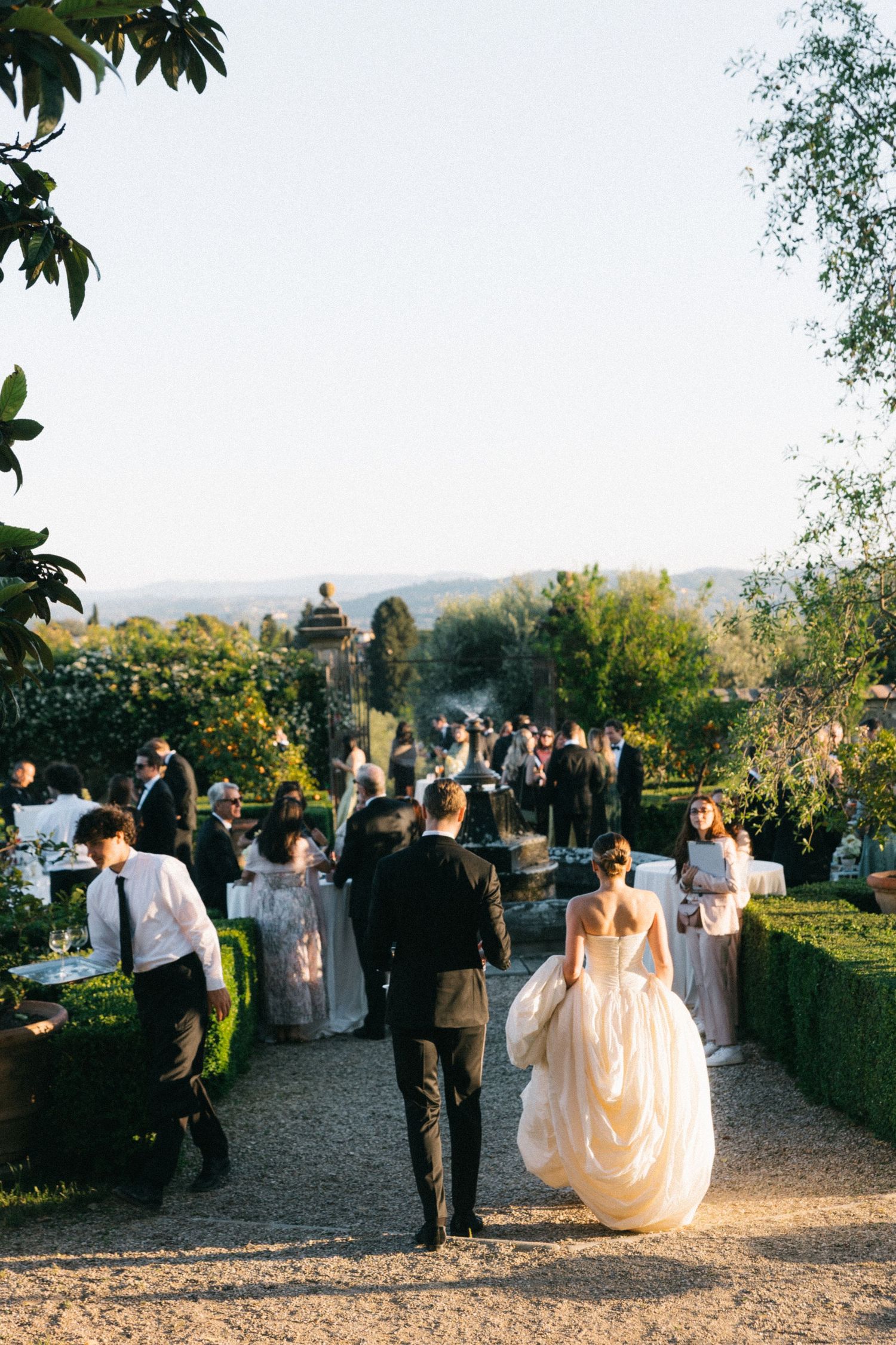 Bride and groom walk hand-in-hand through an elegant outdoor garden wedding reception at golden hour.