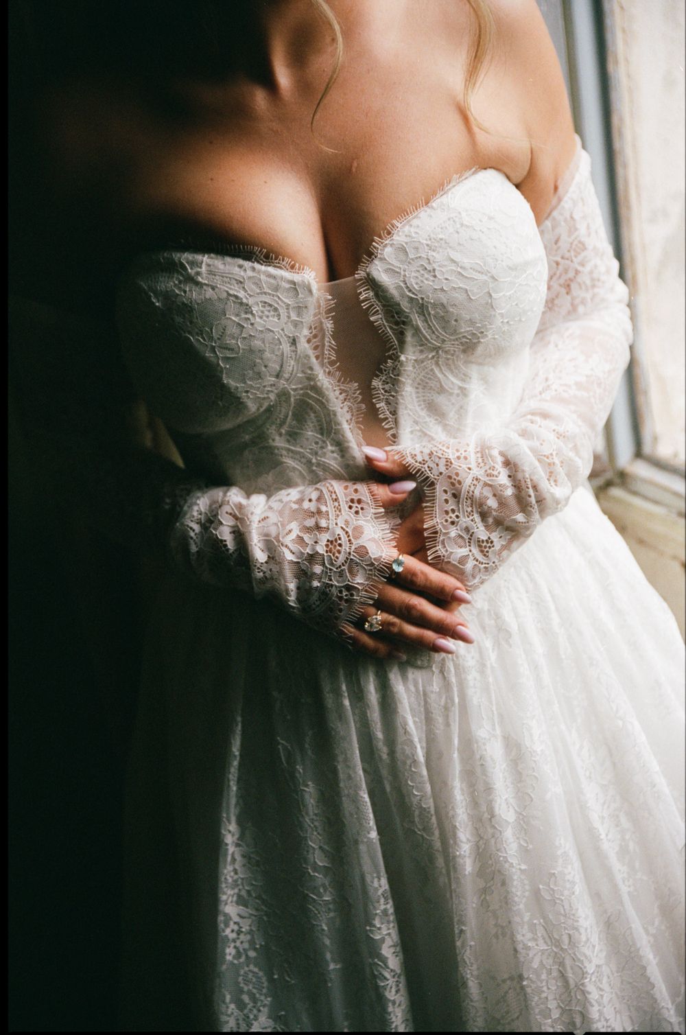 Close-up black and white photo of bride in off-shoulder lace wedding gown by window.