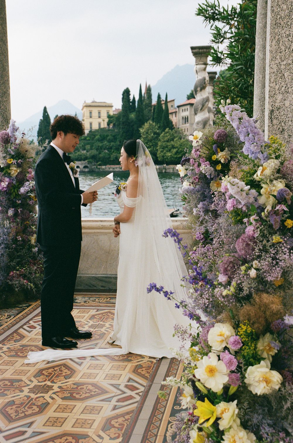 Couple exchanging vows at an outdoor wedding ceremony surrounded by lush purple and yellow floral arrangements by a lakeside villa.