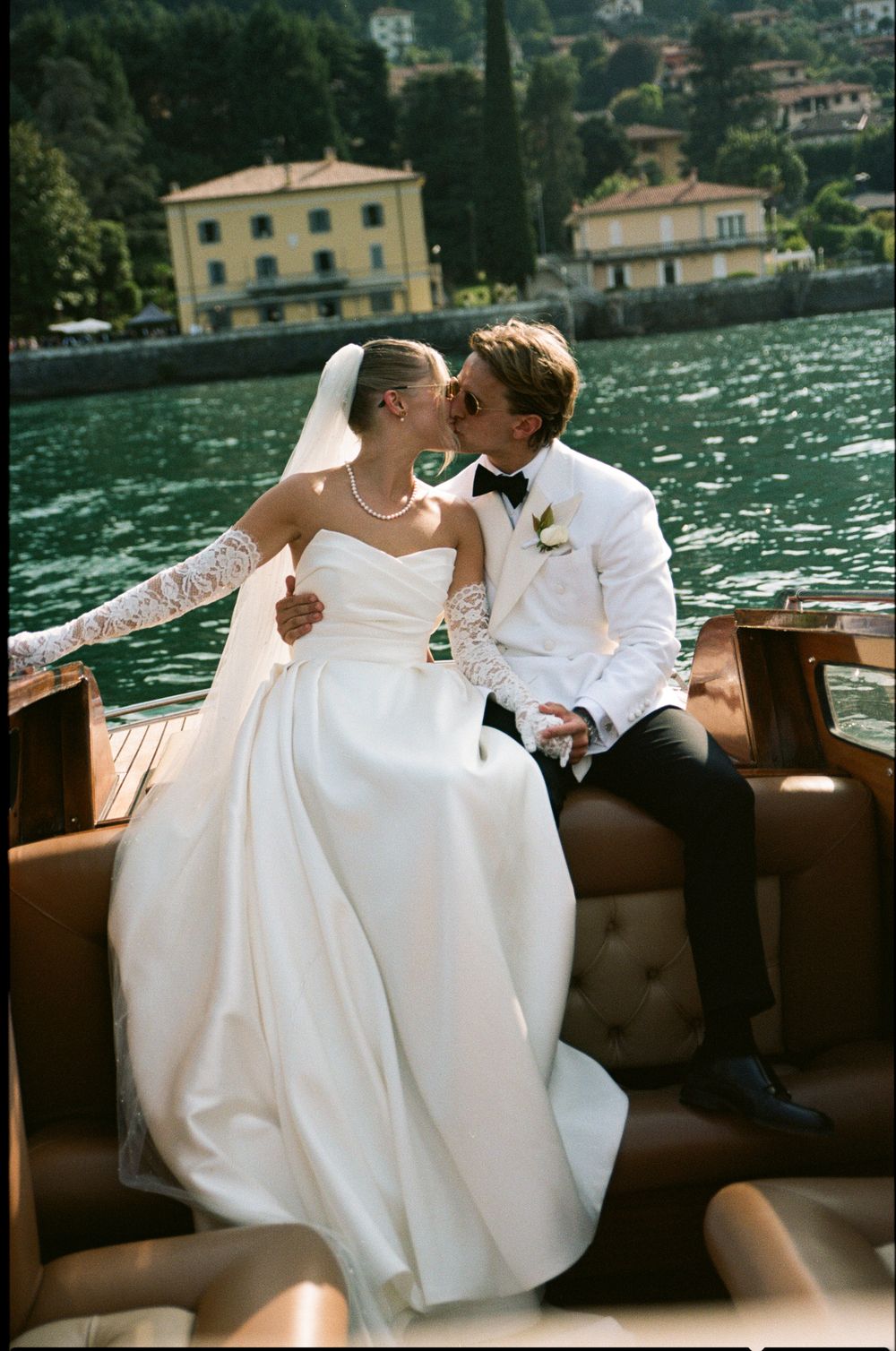 Bride and groom kissing on a wooden boat with scenic Lake Como waterfront behind them.