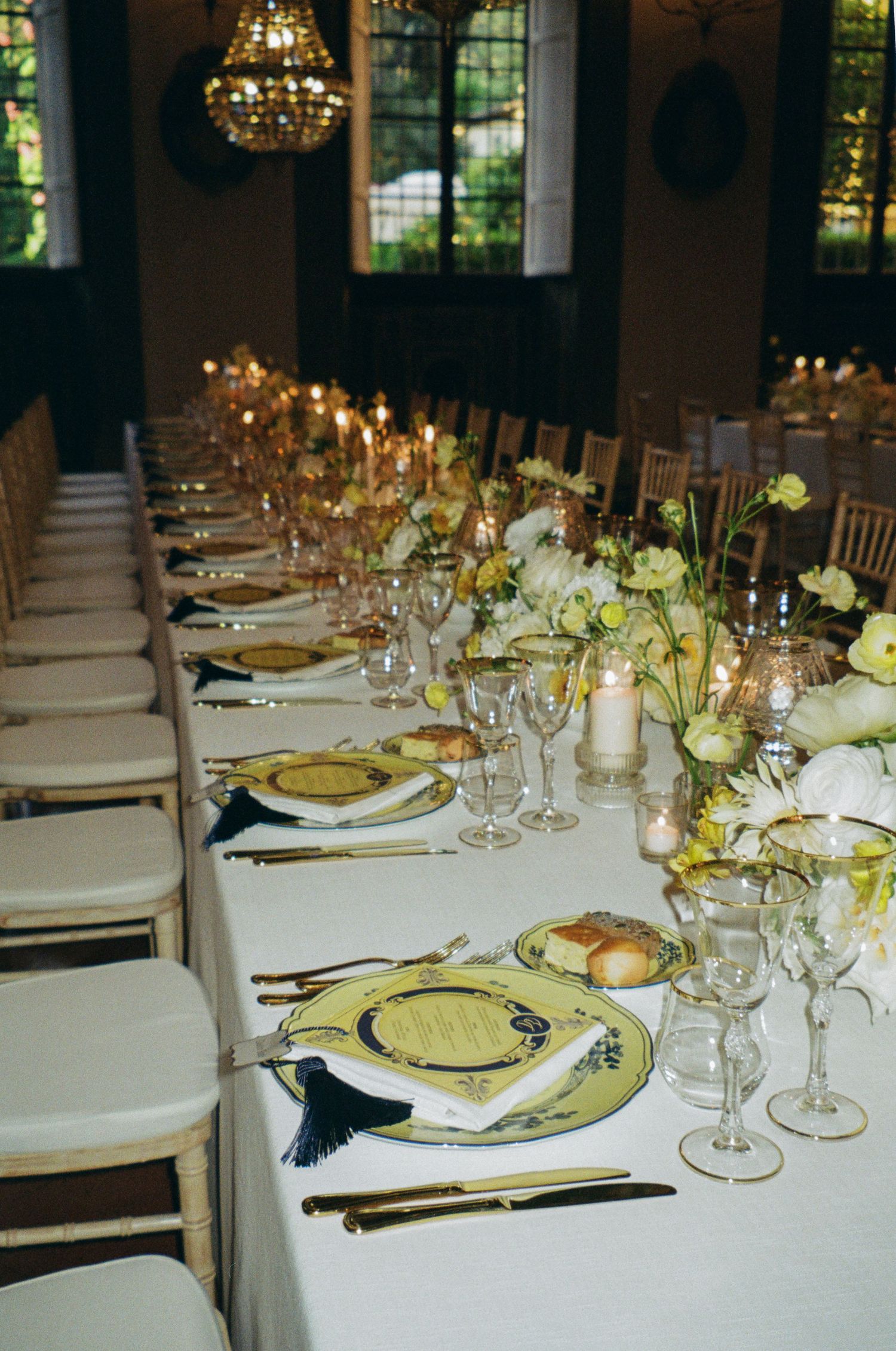 Elegant wedding reception table with gold-rimmed plates, tassel napkins, candles, and white floral centerpieces.