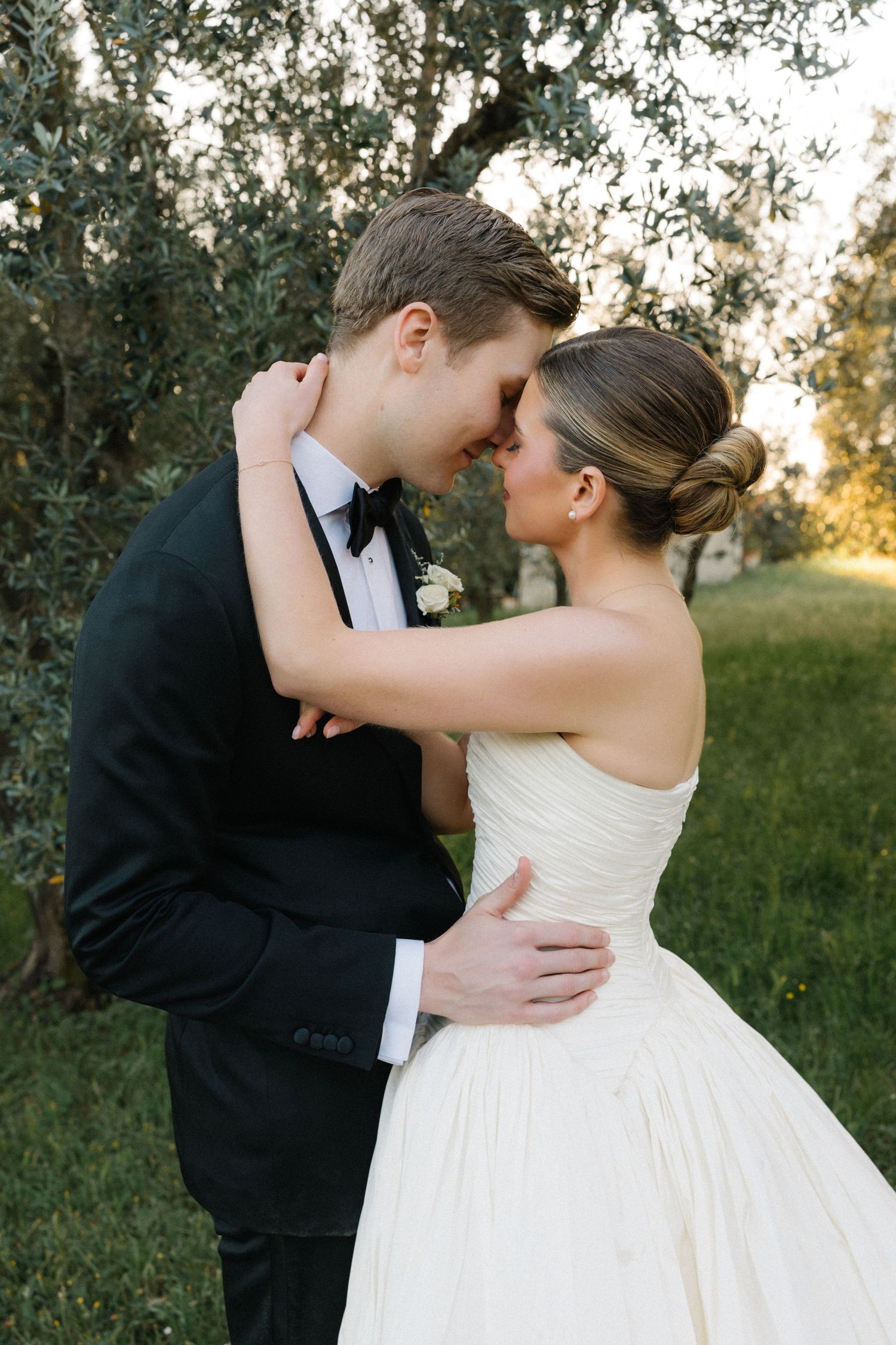 Bride and groom sharing romantic forehead touch embrace among olive trees in golden hour light.