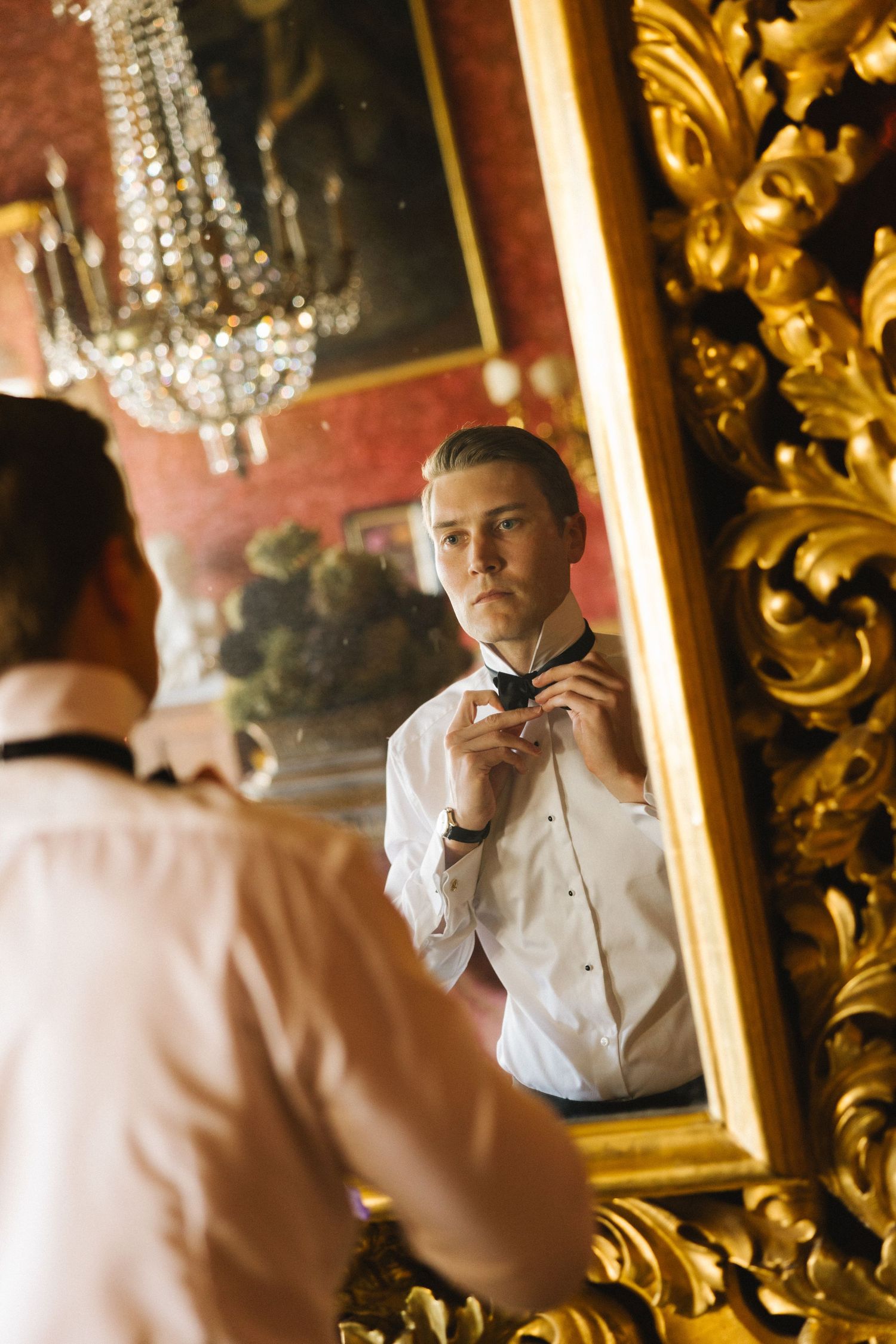 Groom adjusting bow tie in ornate gold-framed mirror with crystal chandelier reflection in elegant room.