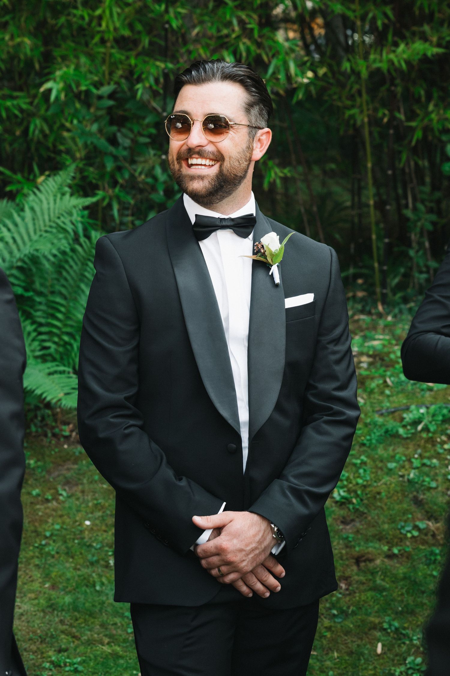 Smiling groom in black tuxedo with bow tie, boutonniere, and round sunglasses standing outdoors at wedding ceremony.