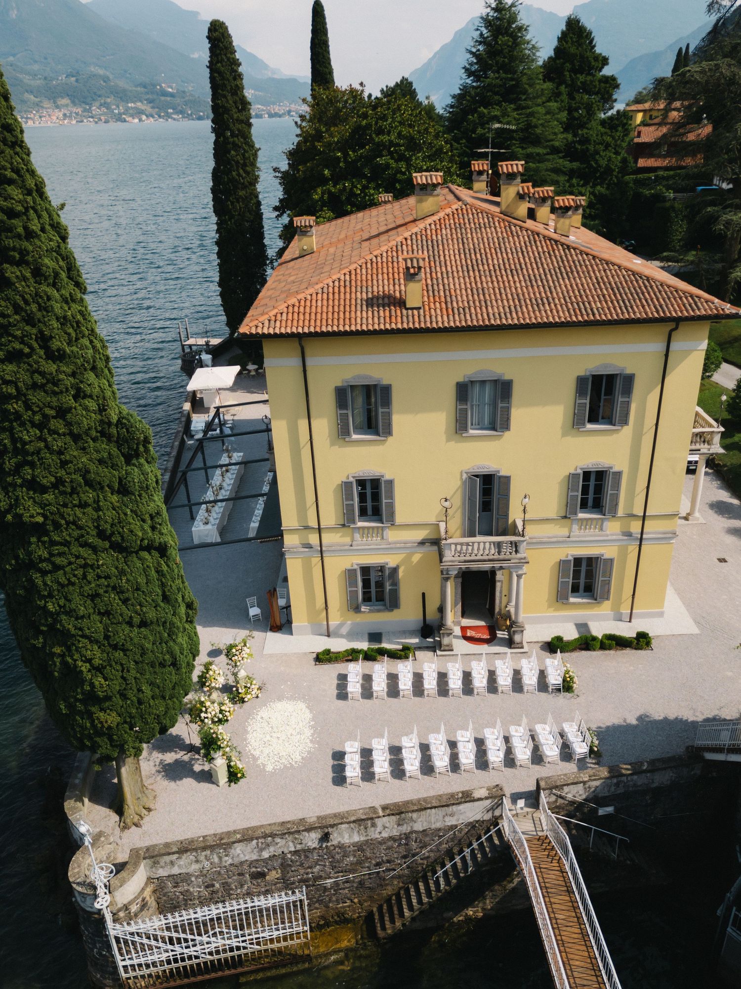Aerial view of a yellow Italian villa with terracotta roof beside Lake Como, surrounded by cypress trees and a stone terrace.