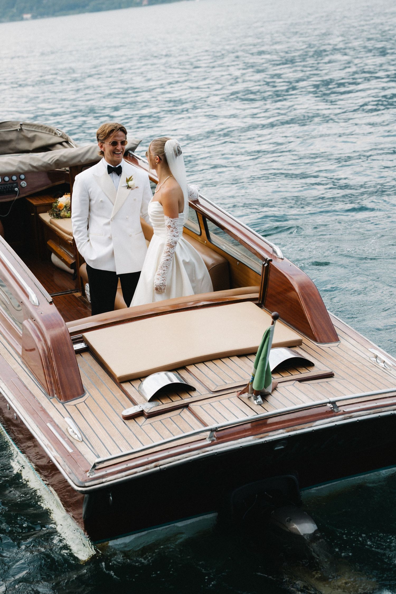 Bride and groom in wedding attire standing on a classic wooden boat on a lake.