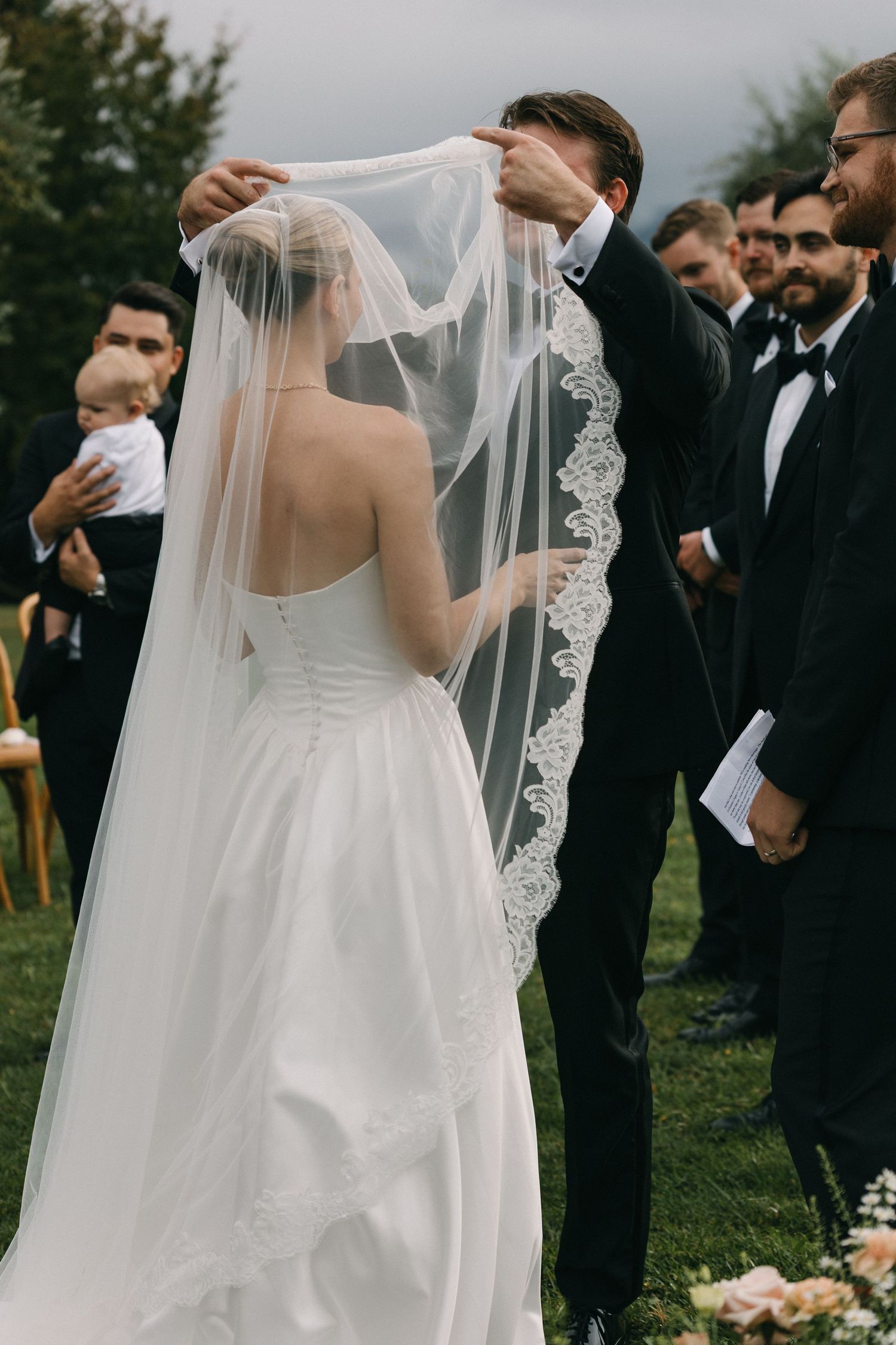 Groom lifting lace-trimmed wedding veil over bride in white gown during outdoor ceremony with groomsmen.