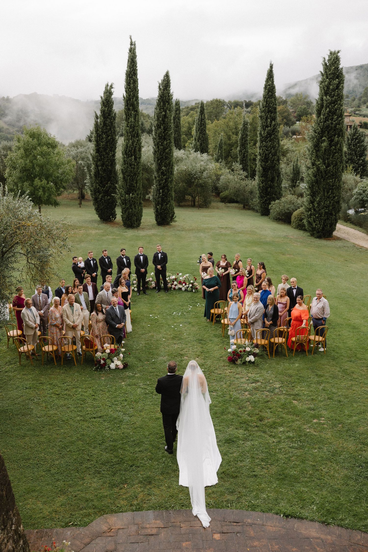 Aerial view of an outdoor wedding ceremony on lush green lawn with cypress trees and misty hills in Tuscany.