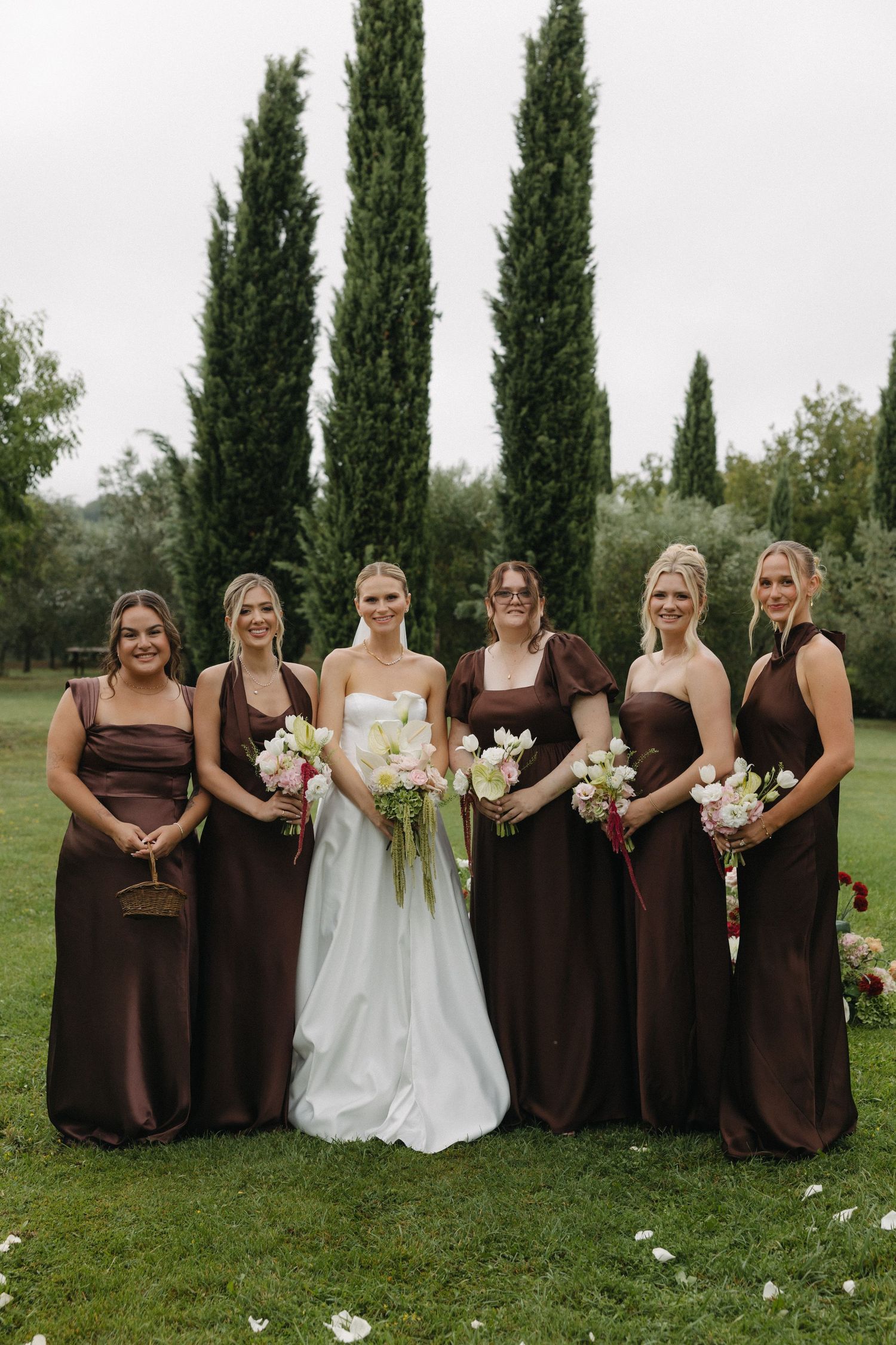 Bride in white gown poses with five bridesmaids in brown satin dresses holding floral bouquets outdoors.