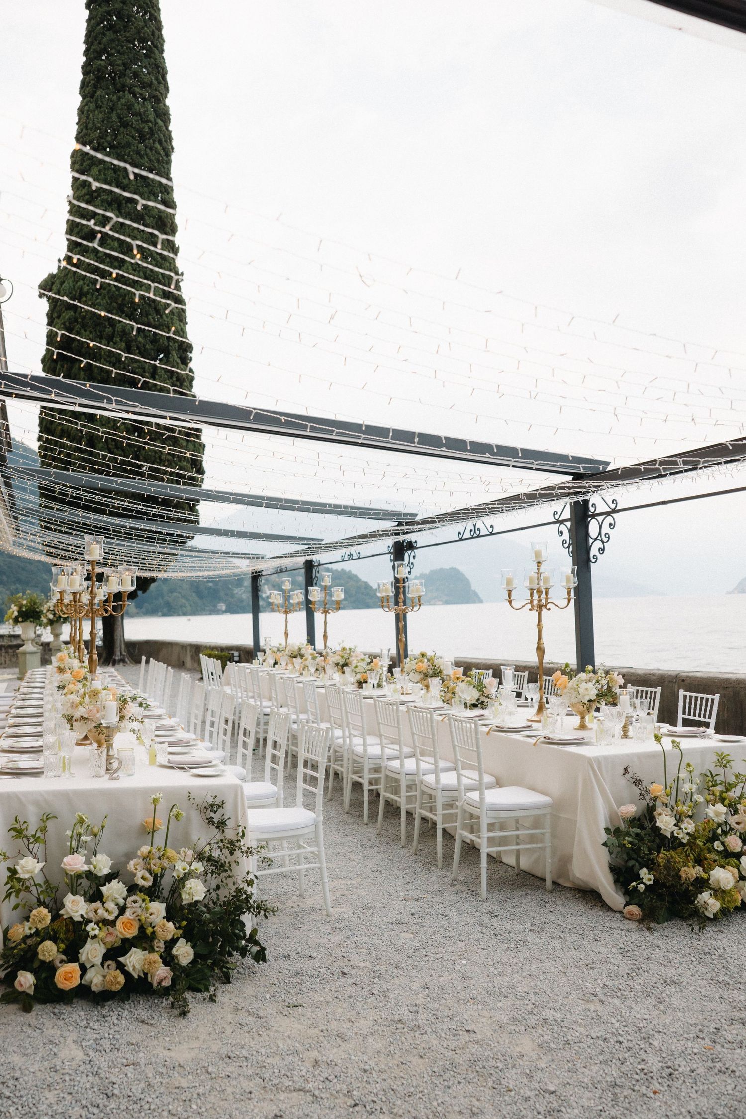 Elegant outdoor wedding reception with white chairs, long tables, floral arrangements, gold candelabras, and a scenic lake view.