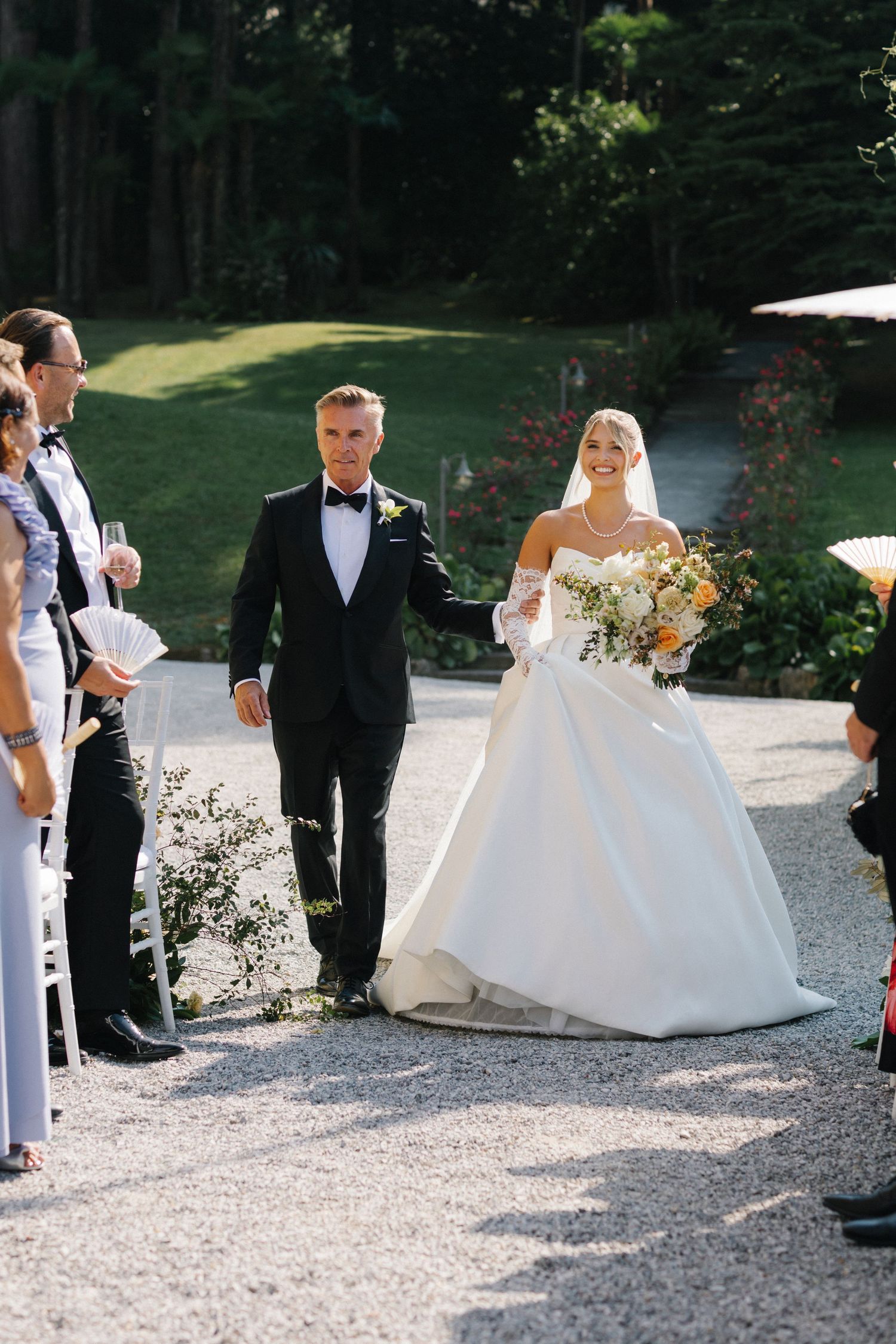 Bride in white ballgown walks down outdoor aisle with escort, holding floral bouquet, guests watching at elegant garden wedding.