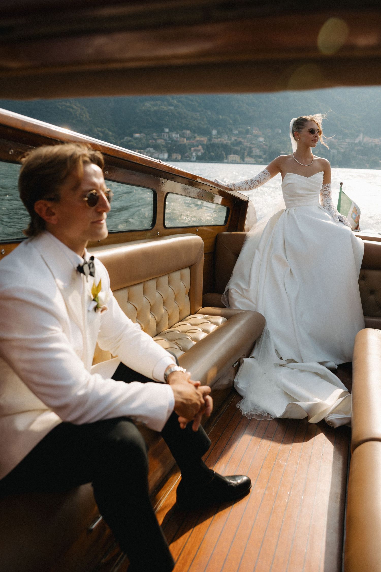 Elegant bride and groom on a luxury wooden boat, wearing wedding attire and sunglasses, with scenic lake views.