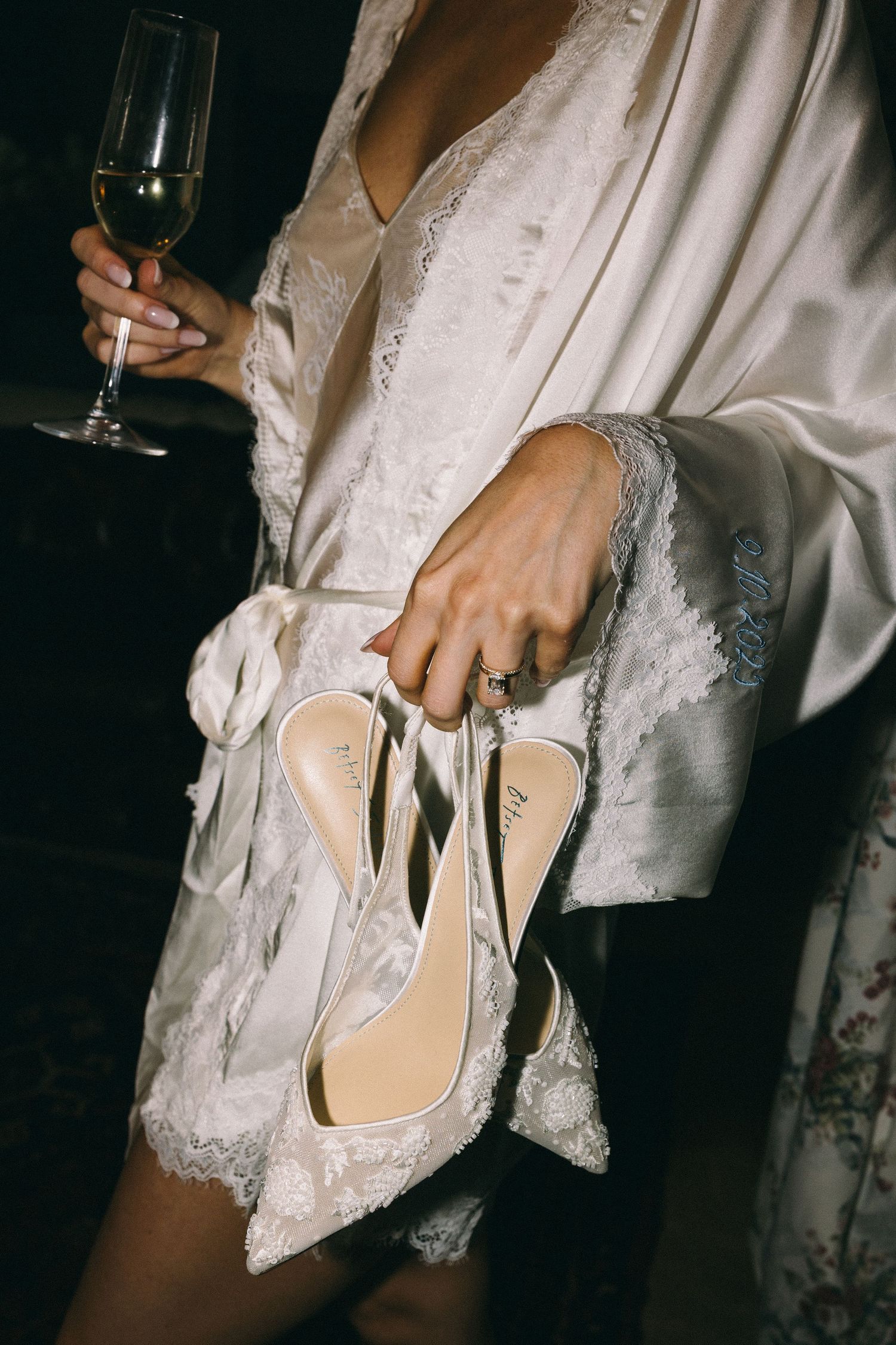 Bride in white lace robe holding champagne glass and elegant white heeled wedding shoes.