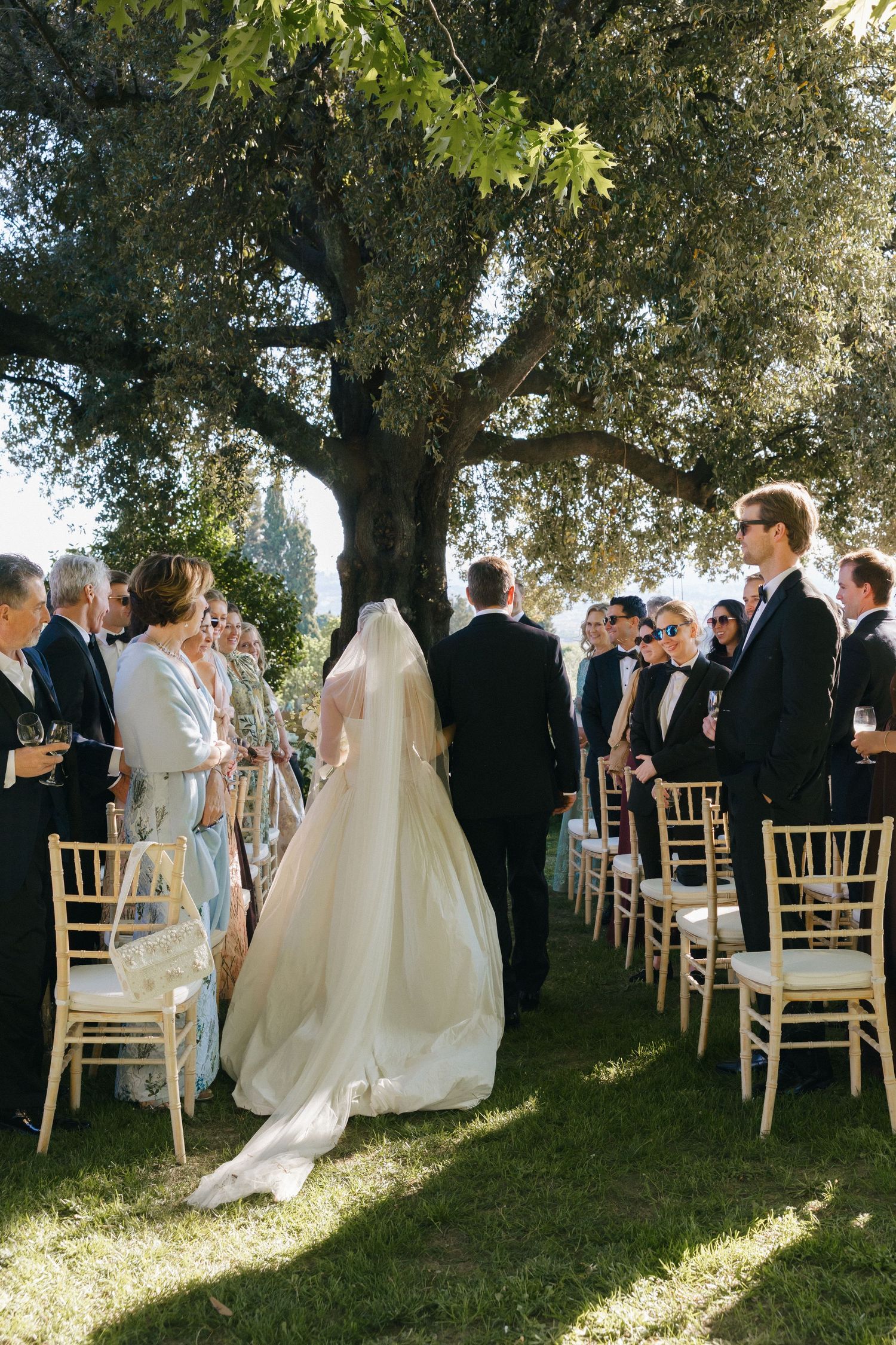 Bride and groom walking down outdoor aisle under large oak tree surrounded by wedding guests in formal attire.
