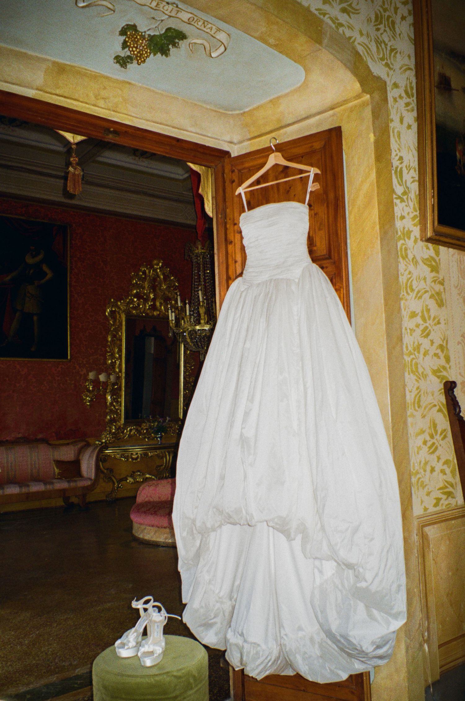 Strapless white wedding ballgown hanging on a hanger in an ornate Italian villa room with heels below.