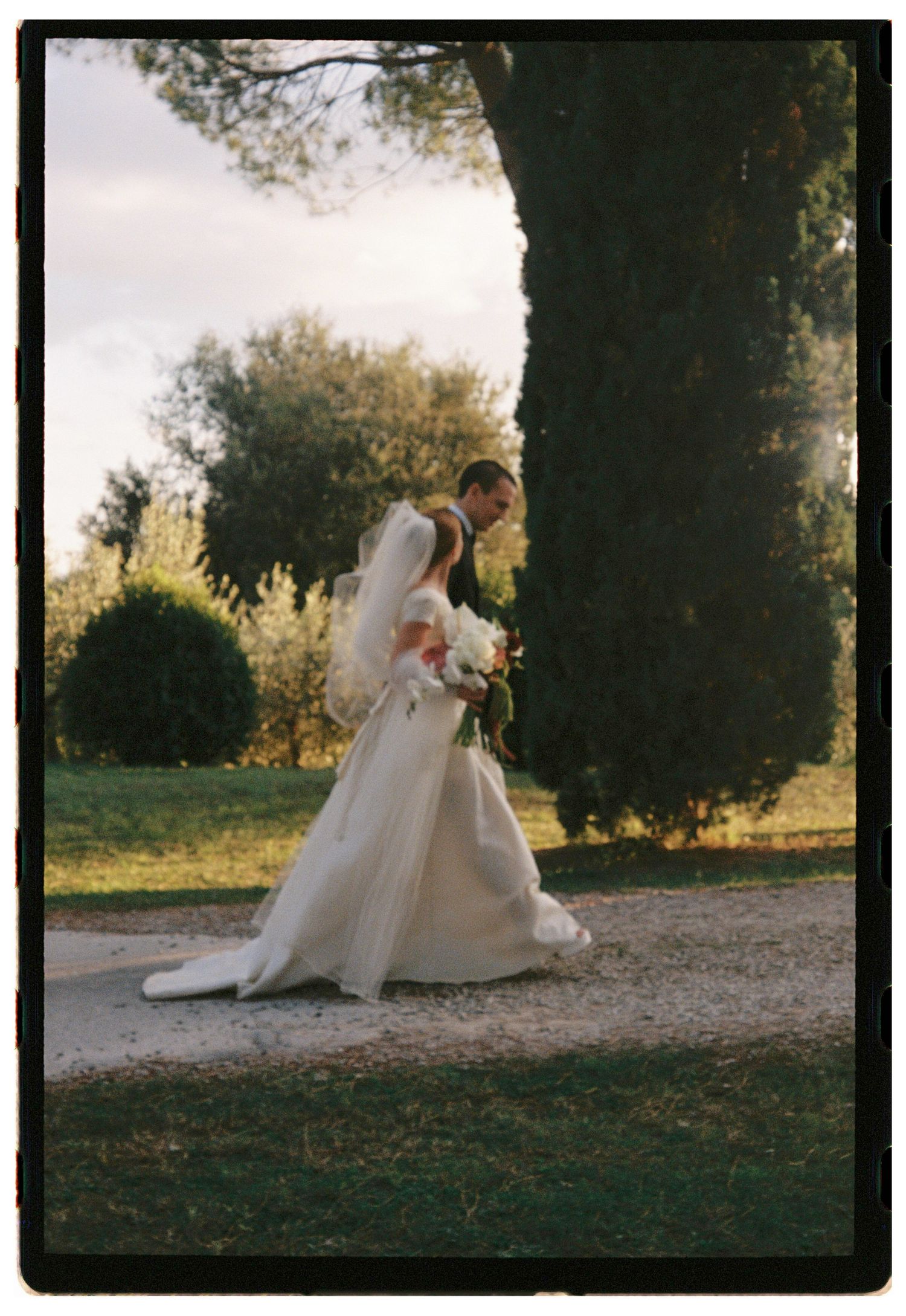 Bride and groom walking together outdoors in a garden setting, bride wearing a white gown with veil and bouquet.
