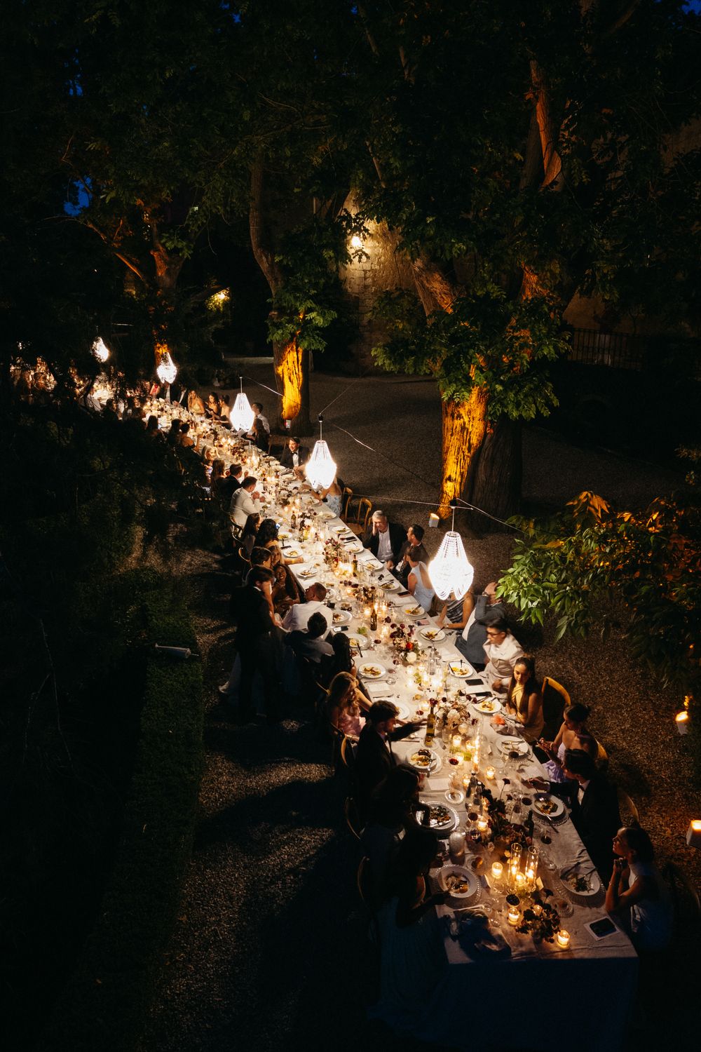 Aerial view of romantic outdoor wedding reception dinner under glowing pendant lights at night.