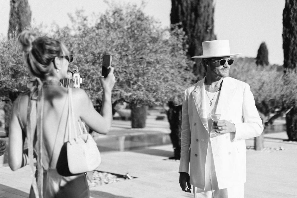 Stylish wedding guest in white suit and hat photographed in black and white outdoor setting.
