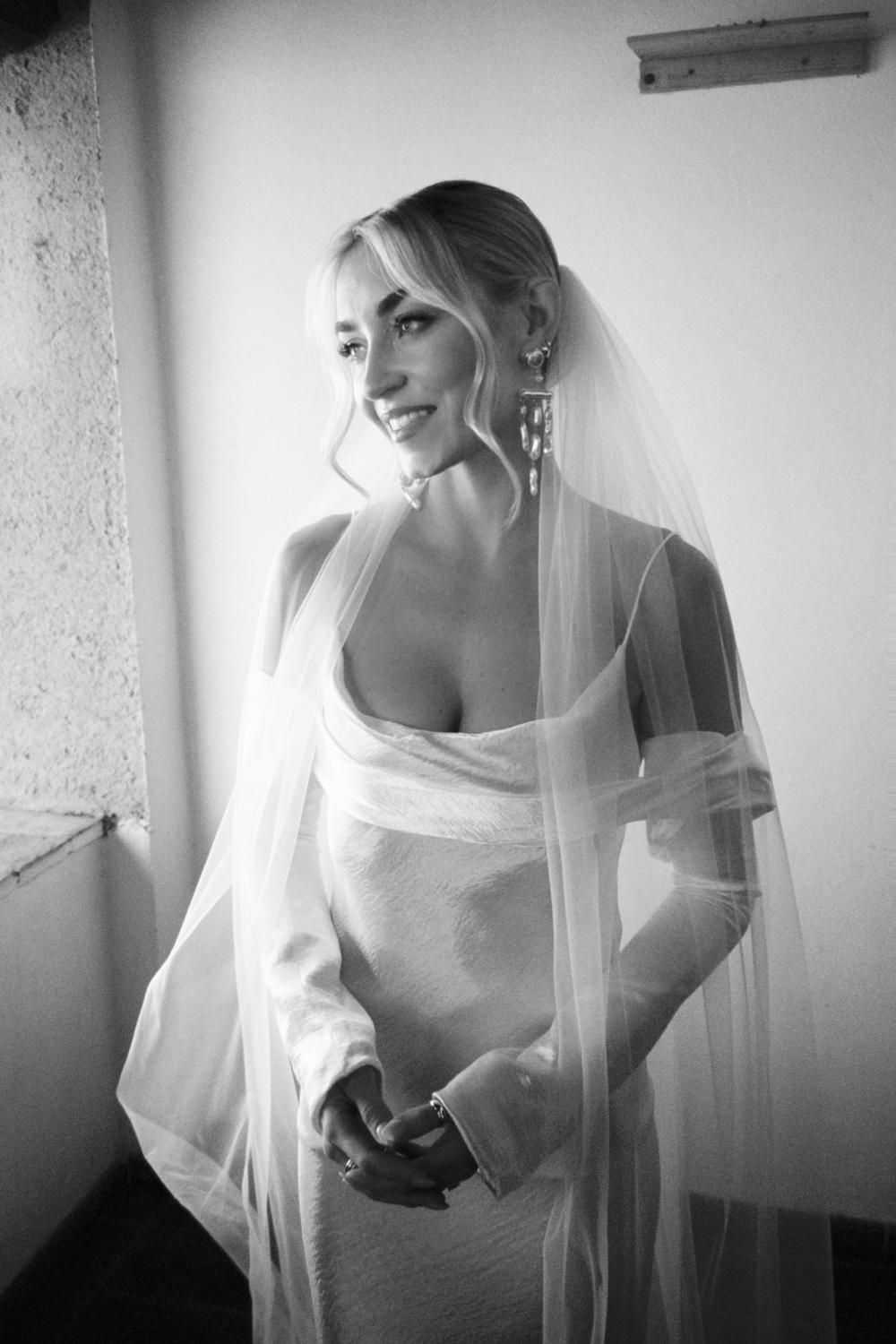 Elegant bride in a fitted white wedding gown and veil smiling softly in a black and white portrait.