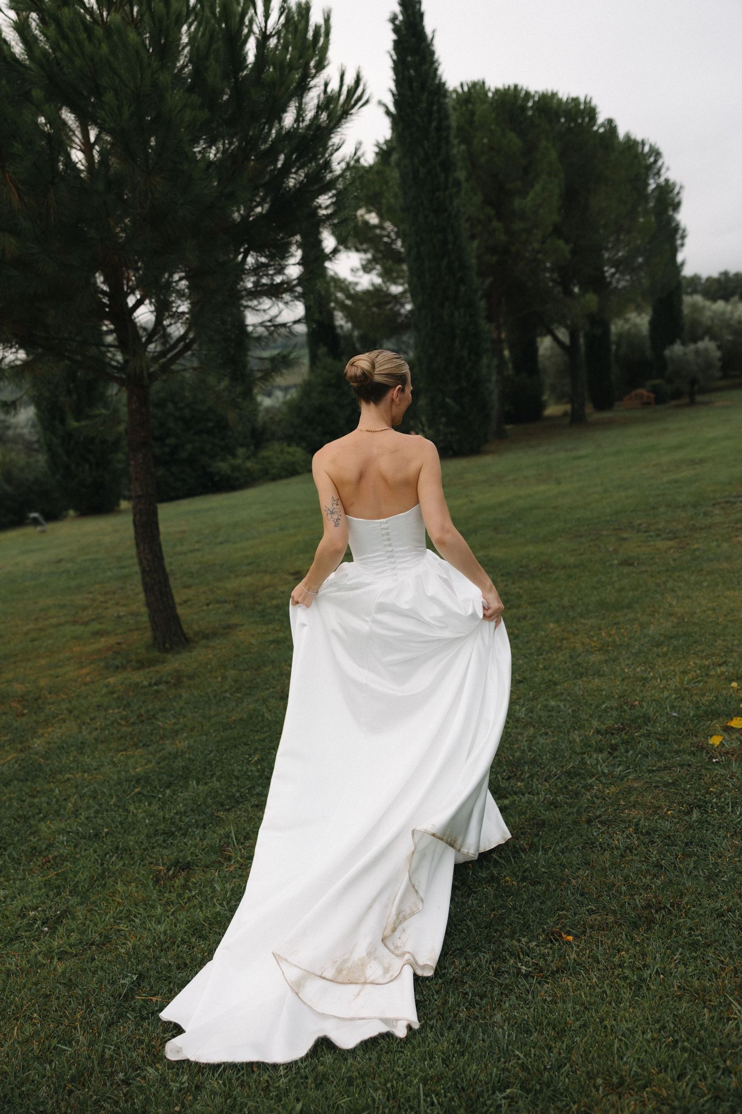 Bride in strapless white gown with train walks through lush green garden surrounded by cypress trees.