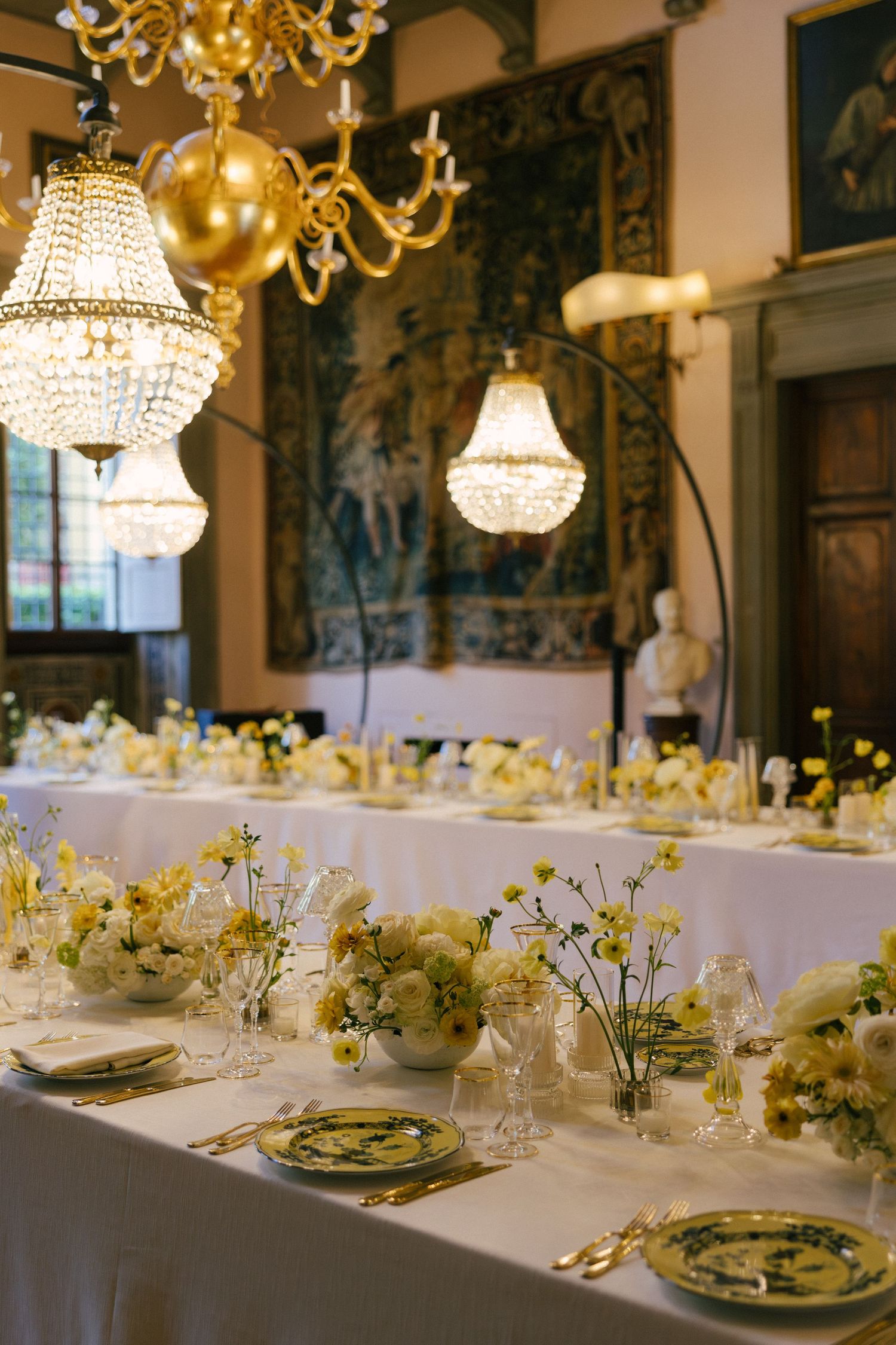 Elegant wedding reception table with white floral arrangements, crystal chandeliers, and gold cutlery in a grand historic venue.