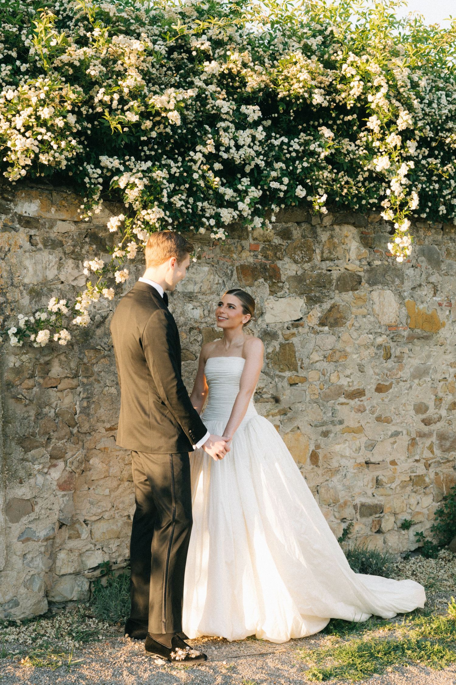 Bride and groom hold hands and gaze at each other beside a rustic stone wall adorned with white blooming flowers.